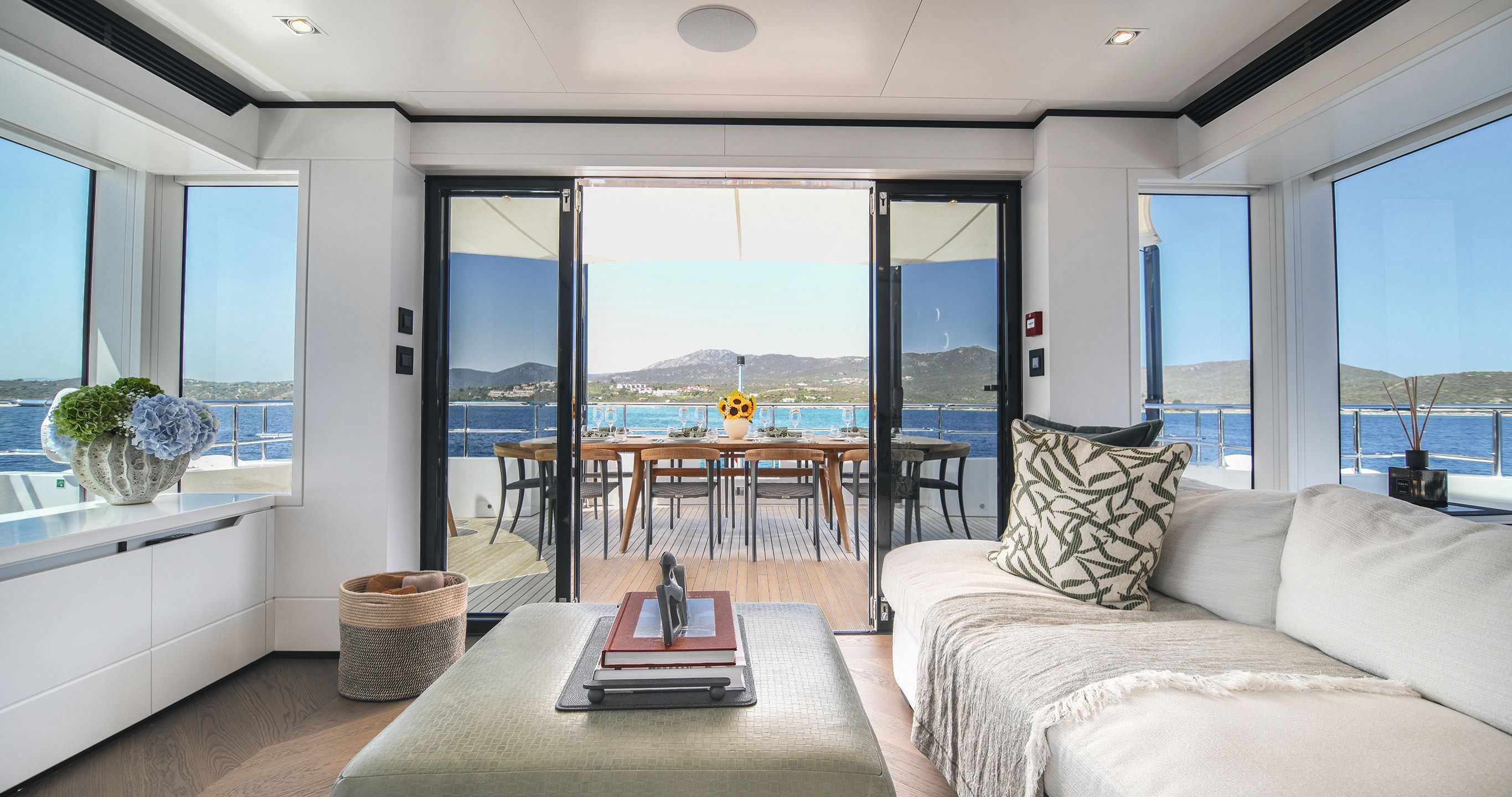 A lounge area with oblong cream sofa facing a table with books on it. There are open sliding doors at the end opening out to the dining table on the deck