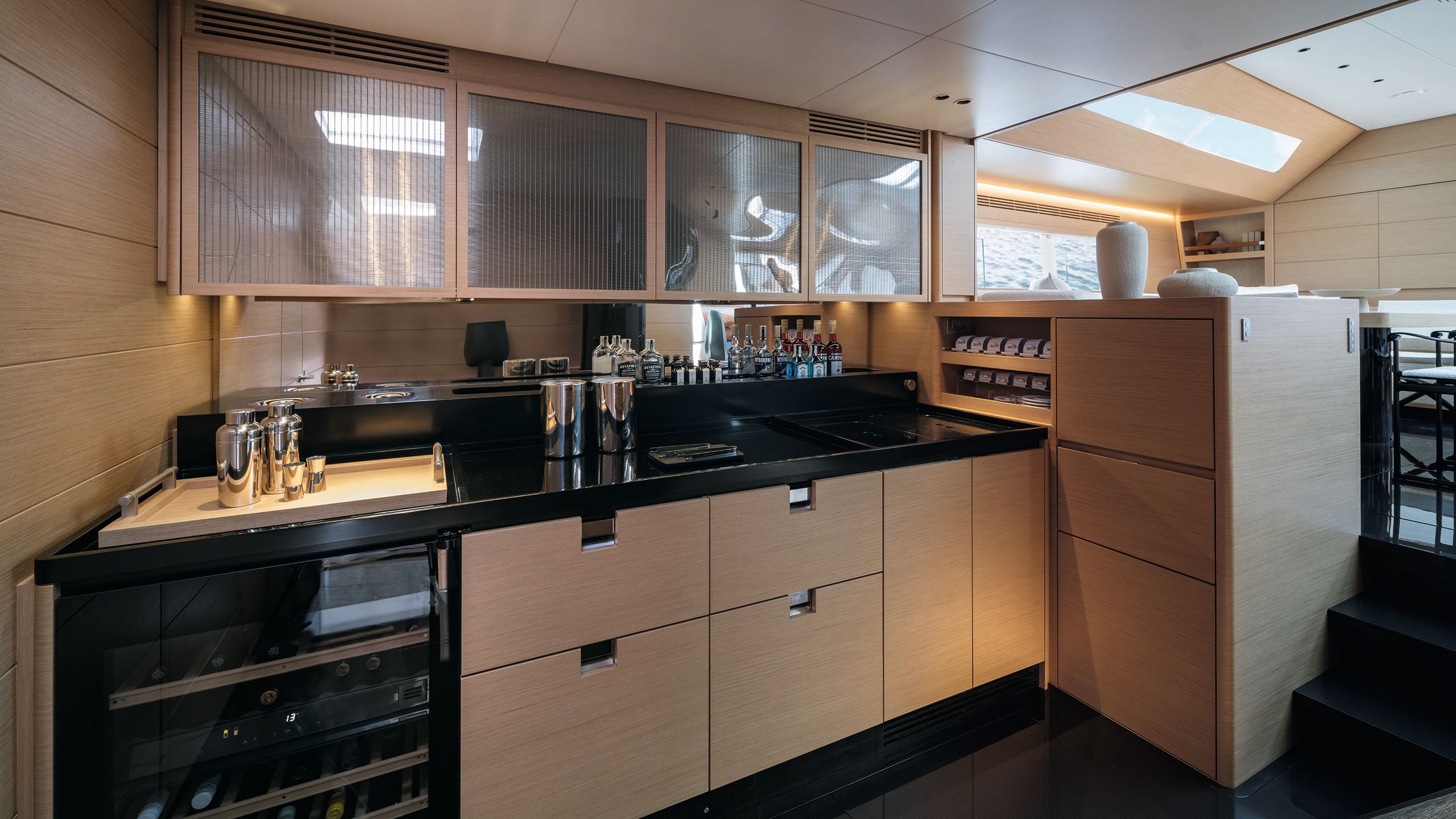 Bar area with cocktail shakers and spirits on a black countertop with oak cupboards at the bottom and ribbed glass cabinets above
