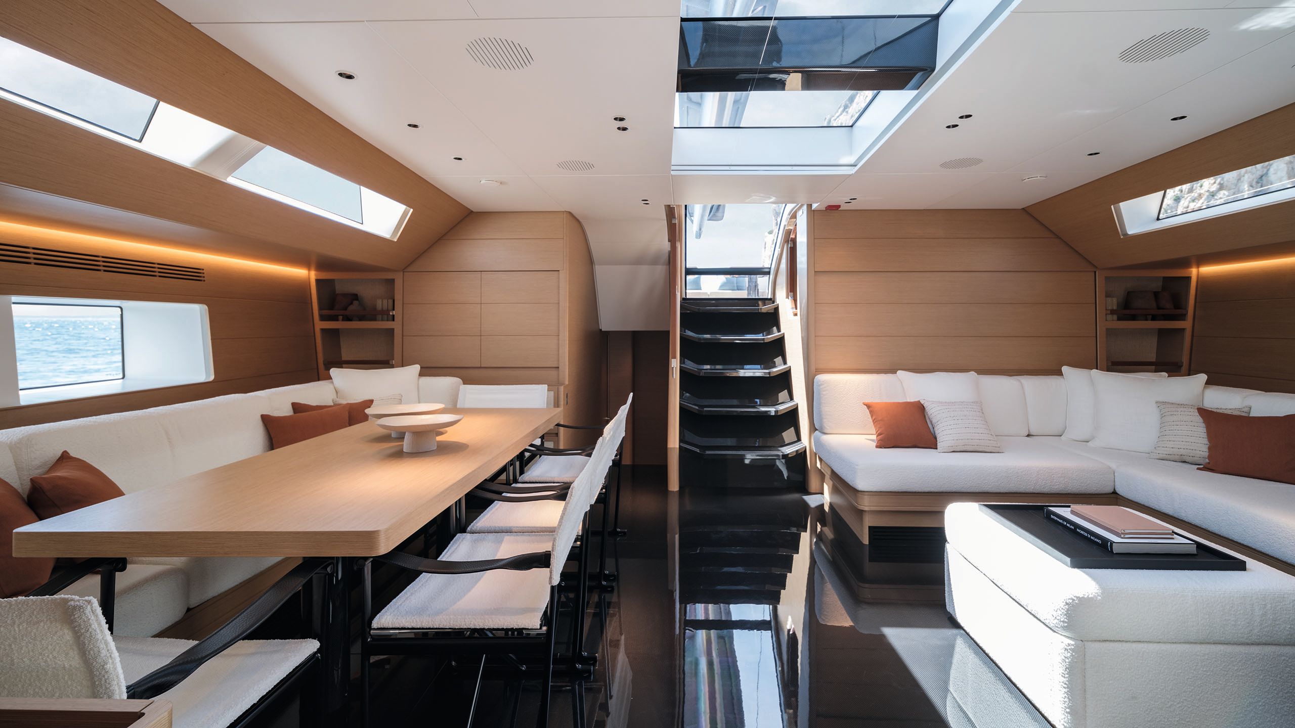 Indoor eating area with table and banquette seating to the left and a u-shaped sofa to the right. The interior is light oak and soft furnishings are white with orange accents