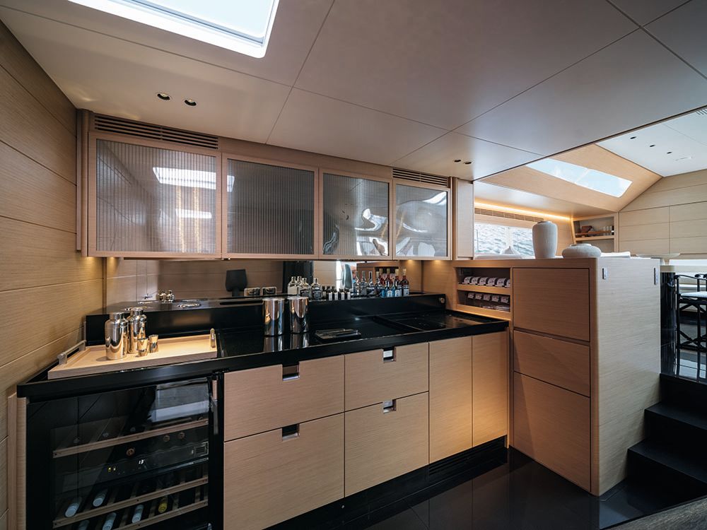 Bar area with cocktail shakers and spirits on a black countertop with oak cupboards at the bottom and ribbed glass cabinets above