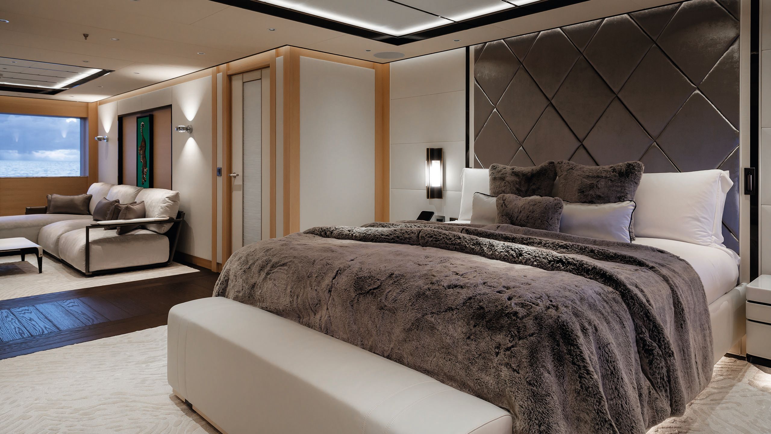 A bed with brown throw and neutral linens. There is a fabric-covered bench at the foot of the bed and to the left, a soft seating area