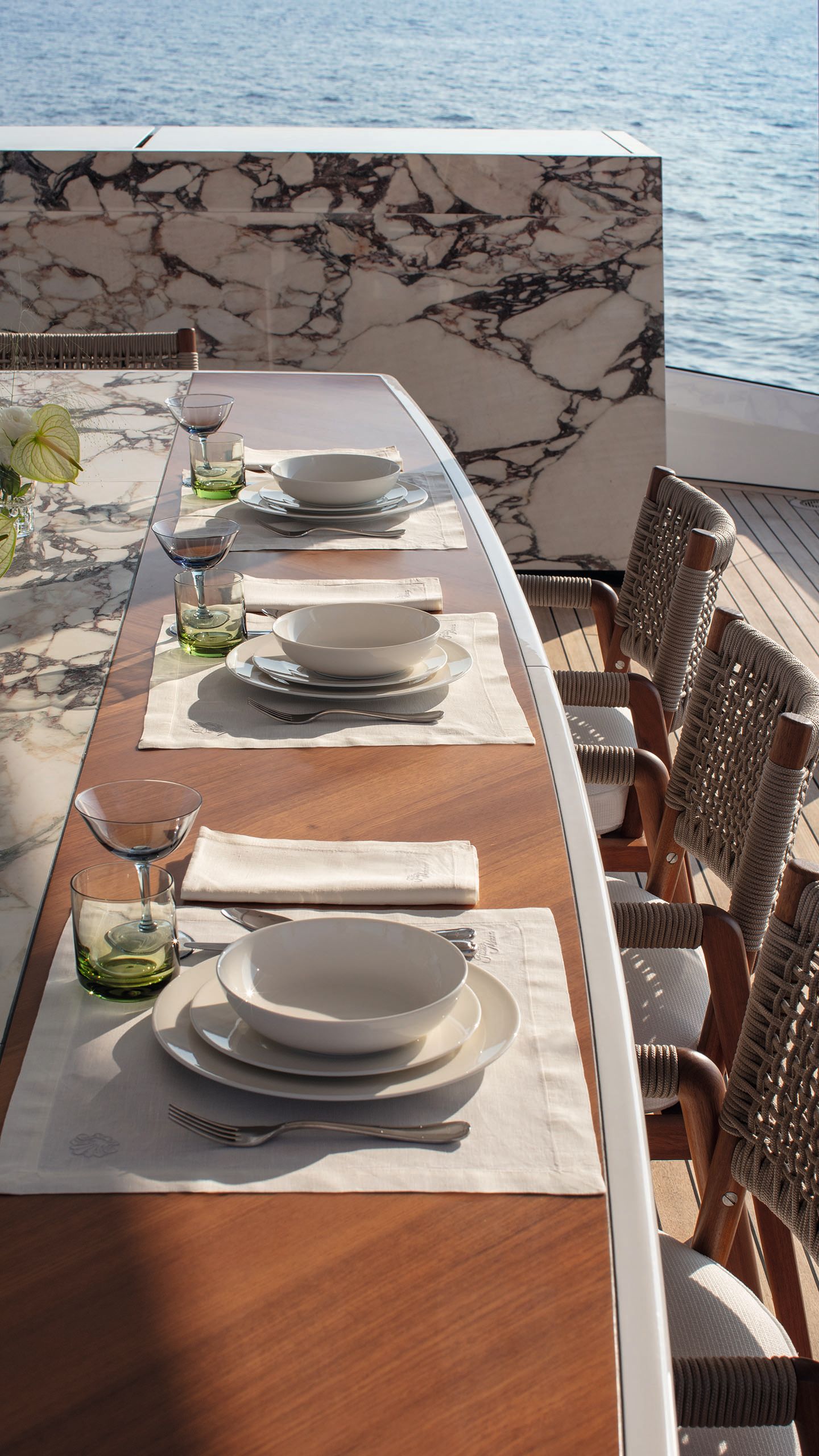 Slightly overhead view of an outdoor marble dining table with teak-coloured inlay. Places are set with white crockery, placemats and napkins
