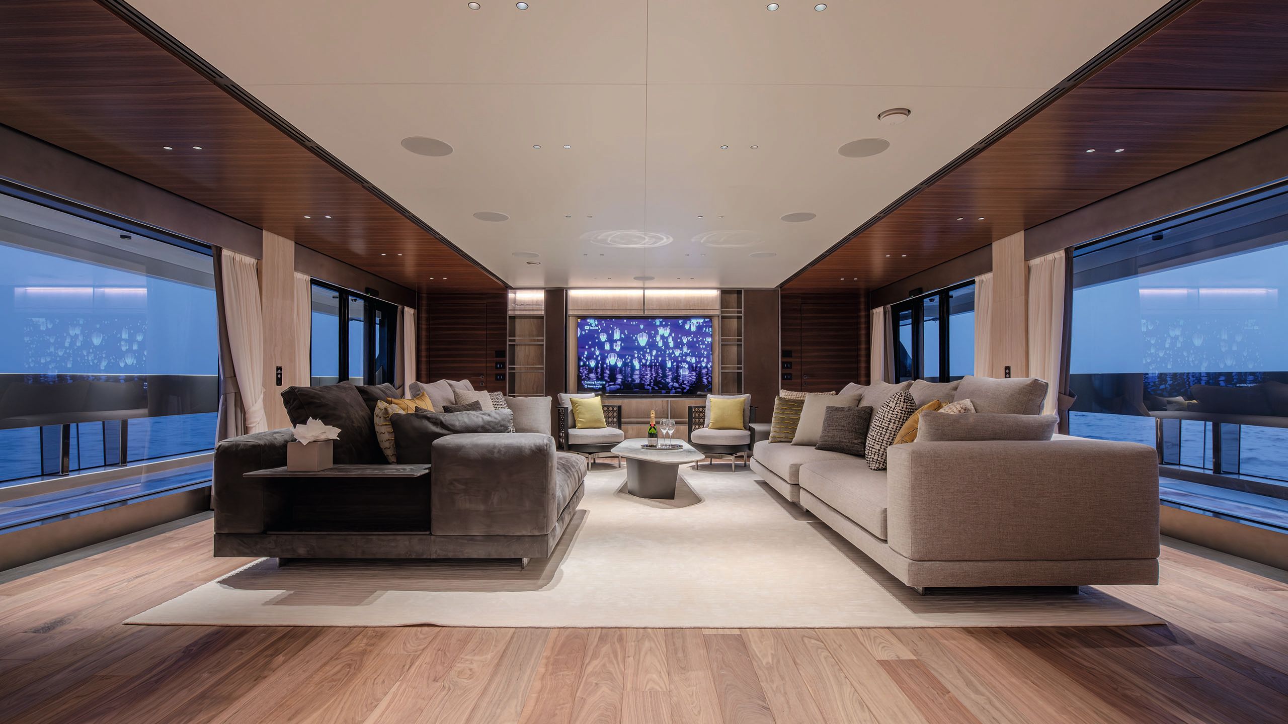 Wide view of the main saloon on Night Fury II. Two large sofas, one beige, one grey, face each other. There are two arm chairs at the end and a coffee table in the centre. In the background is a large wall-mounted TV 