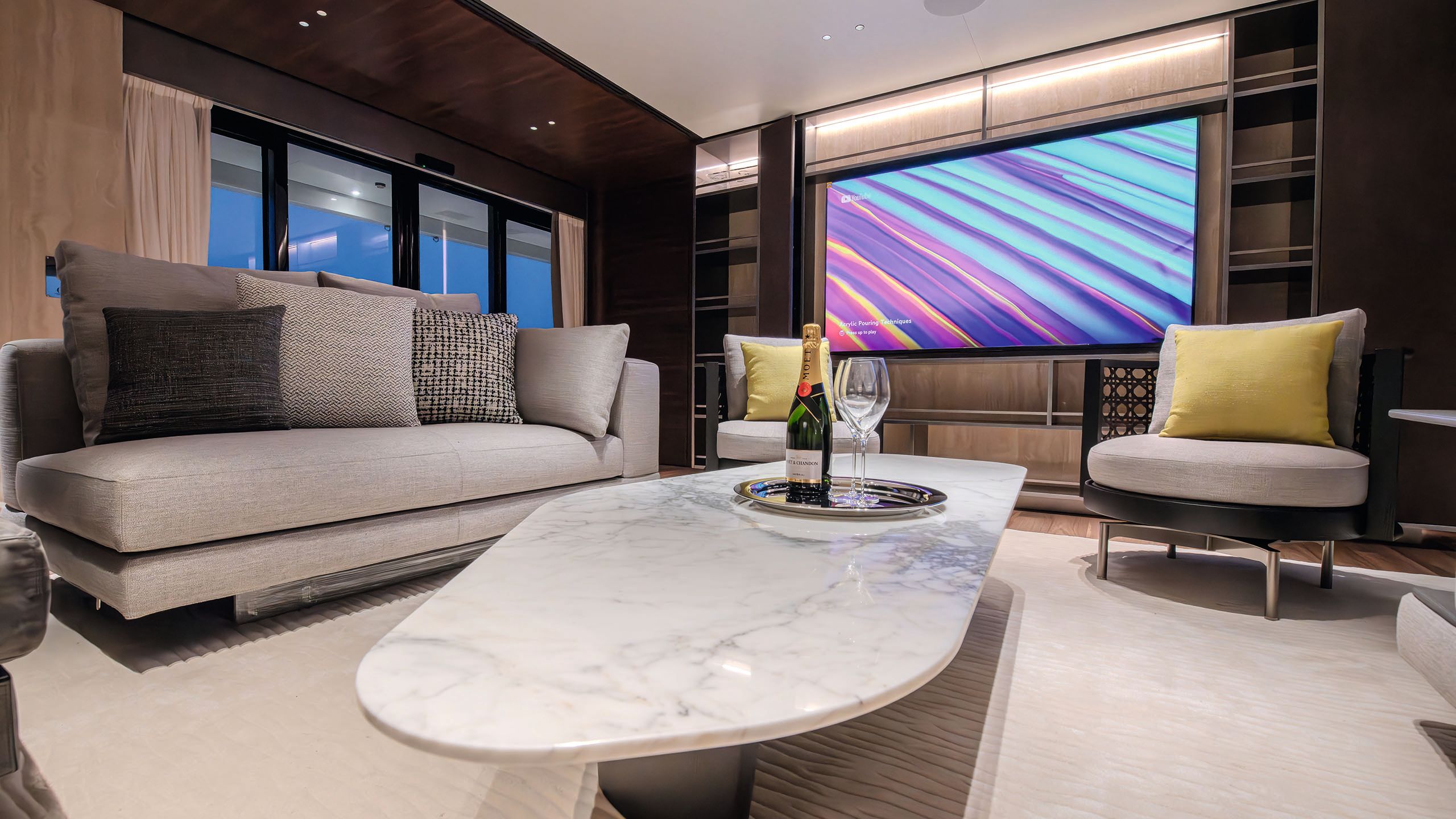 Lounge area on Night Fury II. It has a small beige sofa and matching chairs, large wall-mounted TV and abstract marble coffee table in white. A bottle of Moet and glasses are on a silver tray.