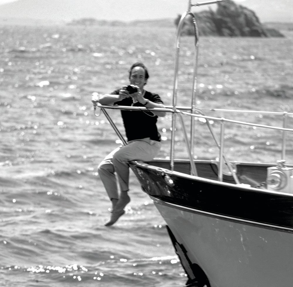 Black and white image of Khan sitting on the edge of a boat with his arms on the railings and his legs dangling over the edge. He's smiling at the camera