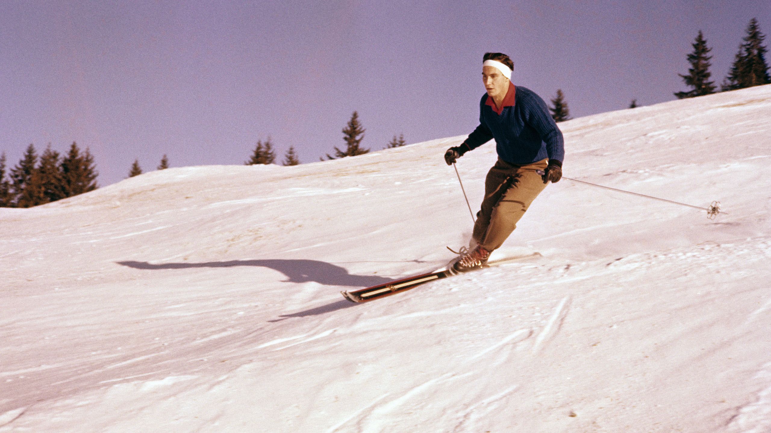 Aga Khan skiing on a snowy slope. He's wearing brown trousers and a blue coat with a white headband round his head. There are trees in the background