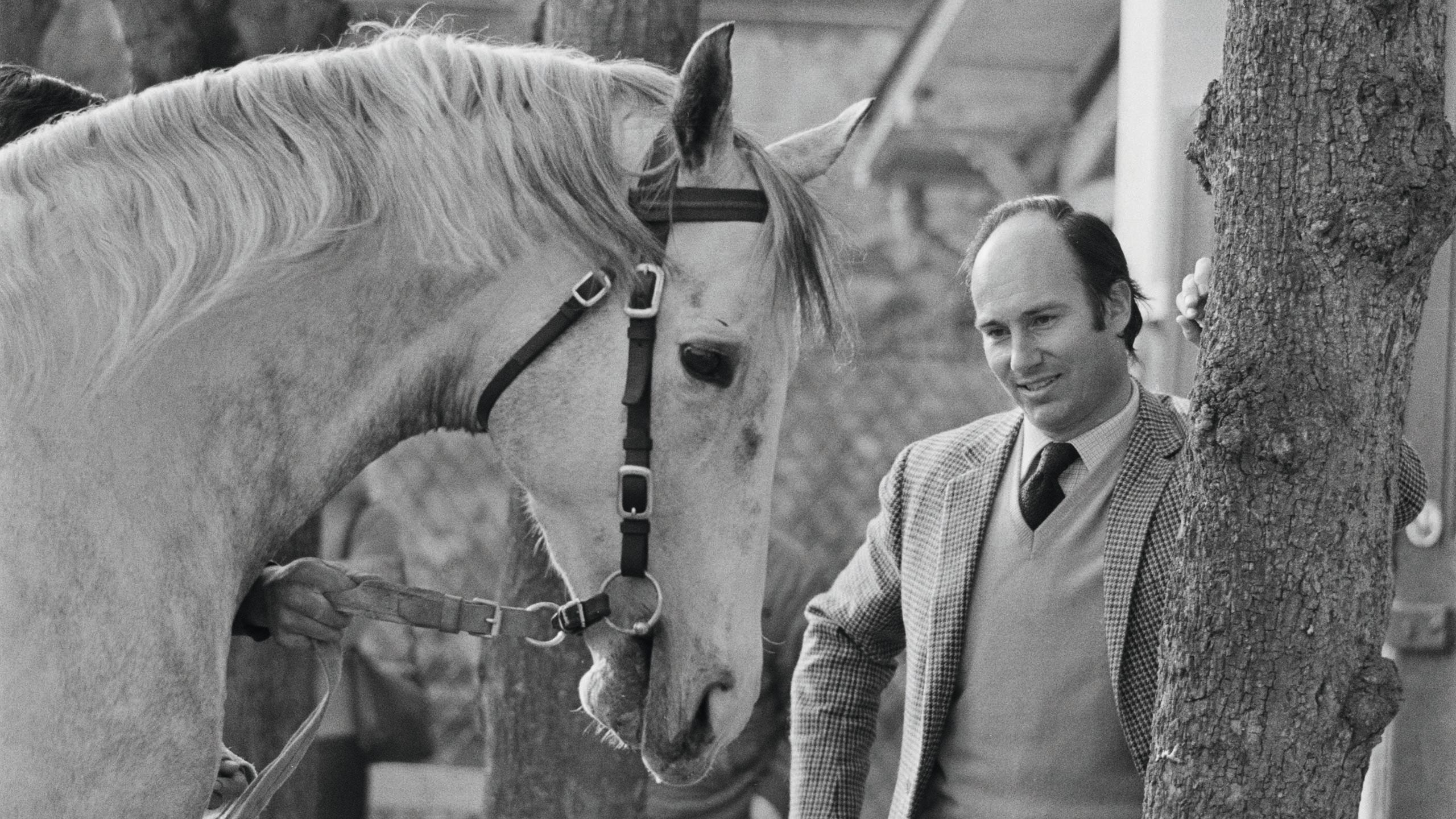 Black and white photo of Khan leaning against a tree trunk next to a white horse