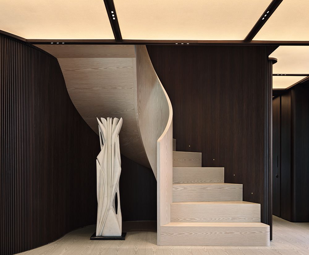 Spiral staircase in neutral colour surrounded by dark panelling and warm lighting overhead