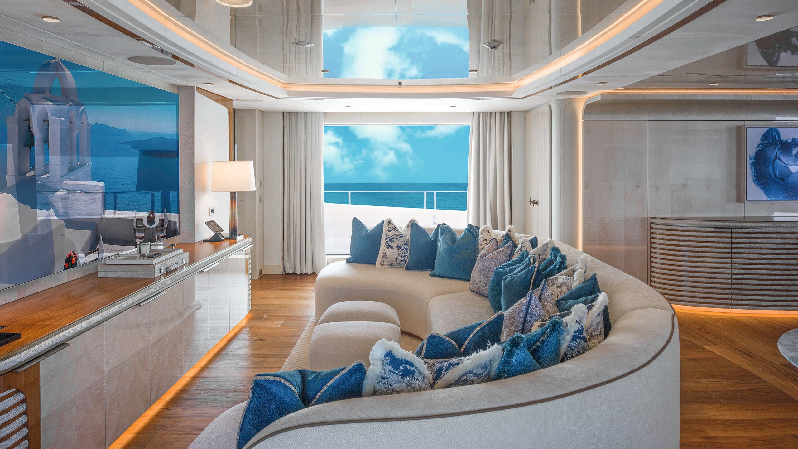 A large crescent-shaped grey sofa facing a wall-mounted TV over a cabinet. There are approx 20 blue and white cushions along the seat pad