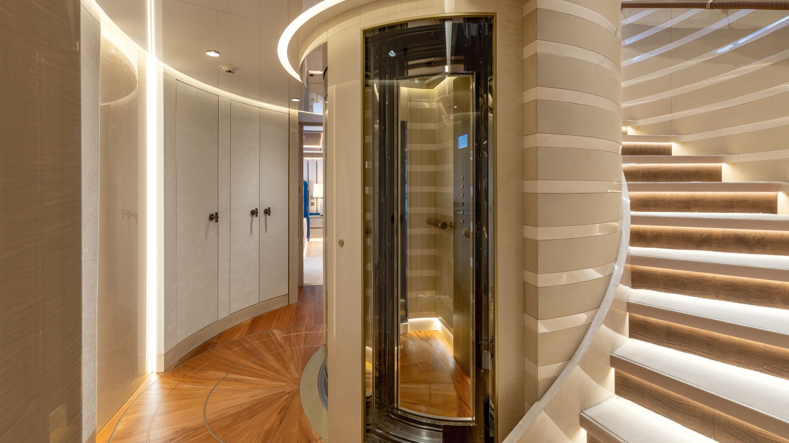 Staircase curving around a glass-front elevator. The floors are teak and the walls/decor a neutral beige/cream