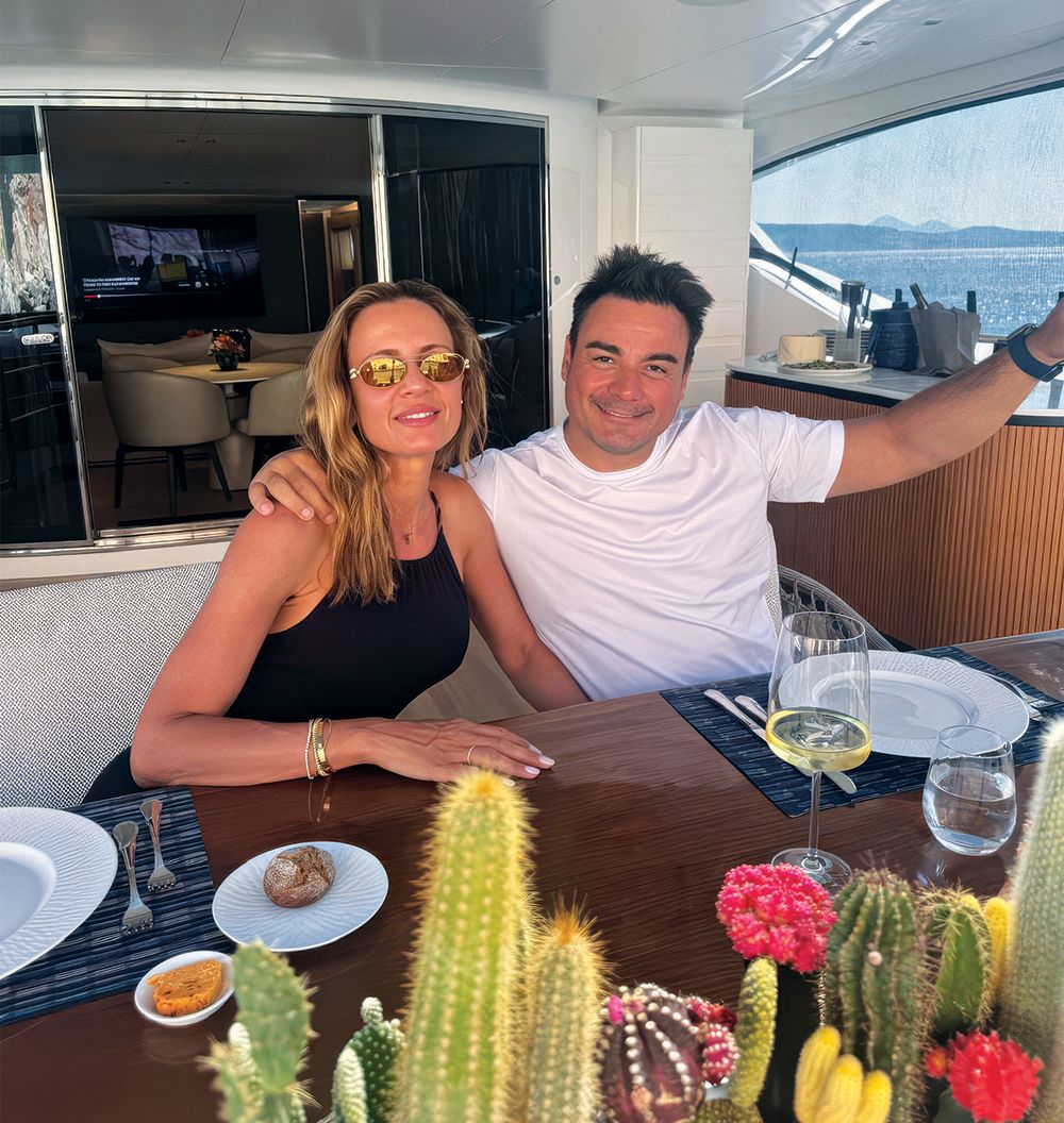 A man and a woman sitting at a table. He has his arm around her and is smiling at the camera, wearing a white T-shirt. She's wearing a black tank-top, sun glasses and is smiling. She also long blonde hair. In the foreground are a selection of cacti