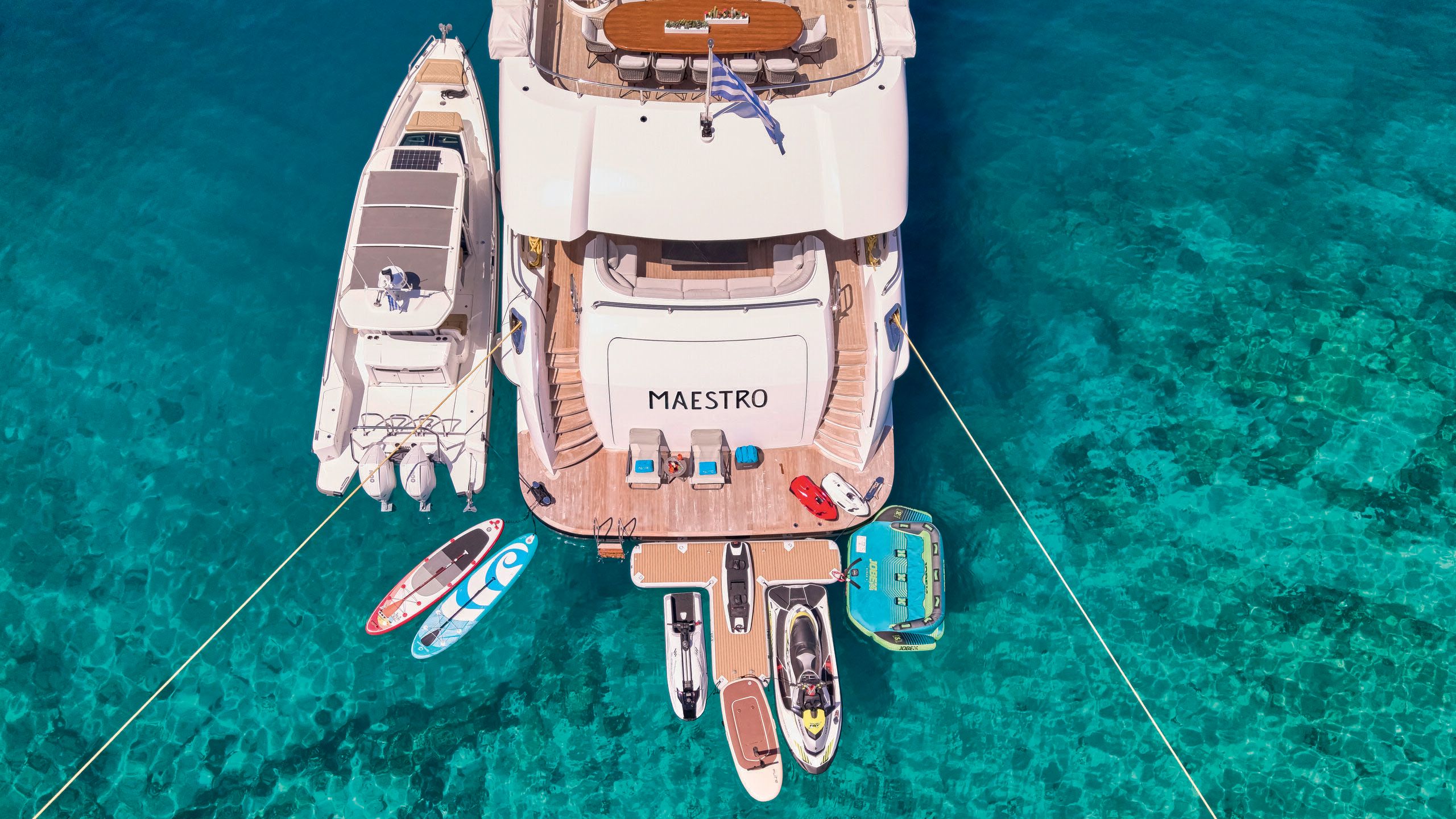 The back of Maestro from above, docked in shallow turquoise waters. There's tenders and paddleboards clustered around the platform