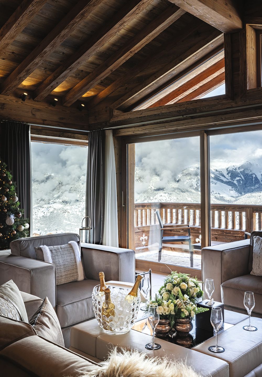 Rustic lounge area of a lodge. There are grey sofas and arm chairs and a large square table in the middle with champagne and flowers in the centre