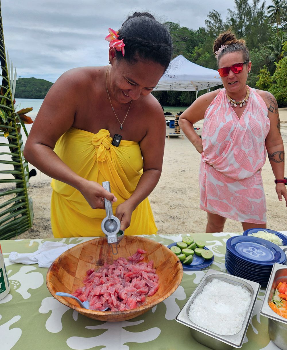 A woman in a yellow sarong dress holding an implement like a citrus presser over a bowl of chopped pink fish chunks. Also on the table is a alrge container of dessicated coconut. A woman behind in watching her