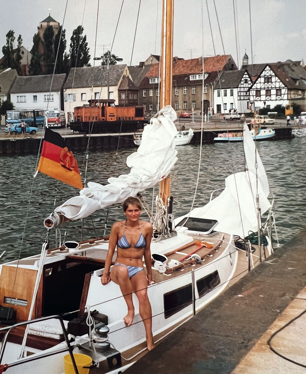 Ramona in a bikini, sitting on a small boat that has a German flag tied to the mast