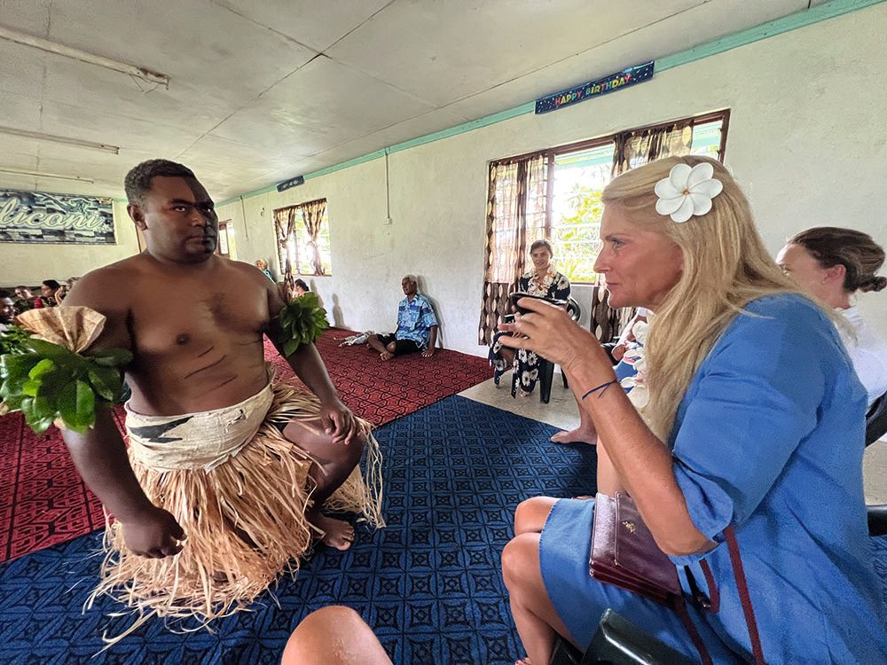 Ramona drinking from a cup. She is wearing a blue dress with a white flower in her hair. A man in traditional dress is kneeling in front of her