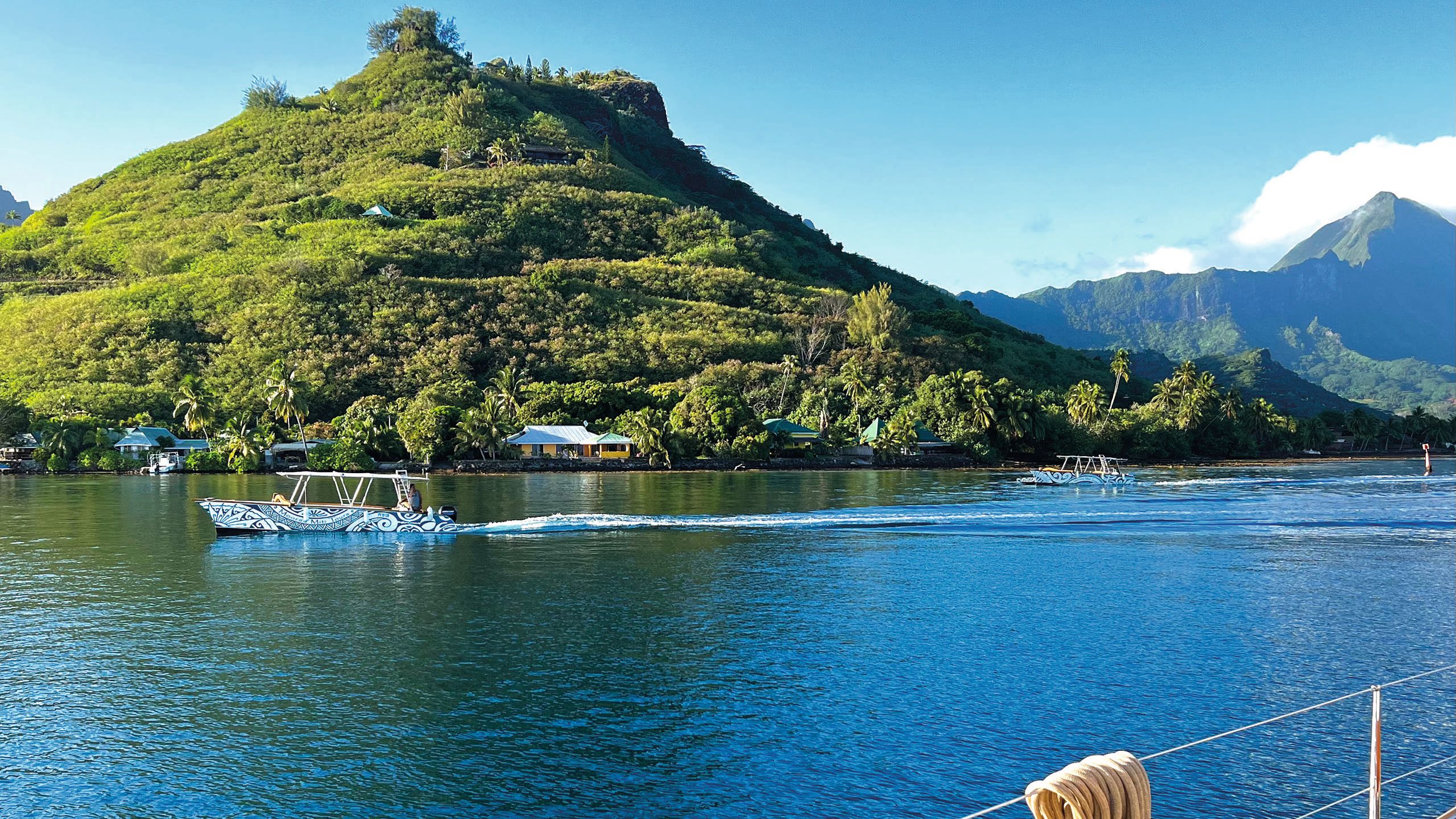 View from the boat looking towards an island covered in lush green vegetation. The water is deep blue and there is a boat in the foreground with a black and white pattern on the side. On the shore are low-level houses