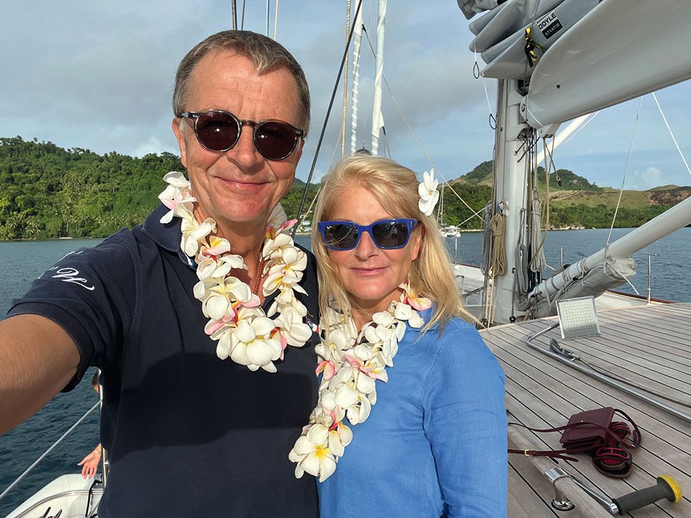Selfie of the Wolles - they are both wearing leis and sunglasses. She is wearing a blue long sleeved top and he is wearing a navy short sleeved top
