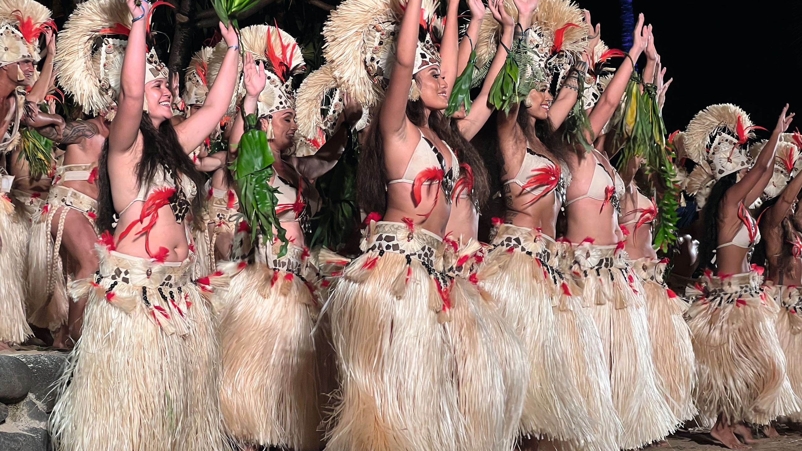 Halva dancers in long skirts made of white grass. They are wearing bikini tops decorated with red feathers and headdresses. Their arms are in the air