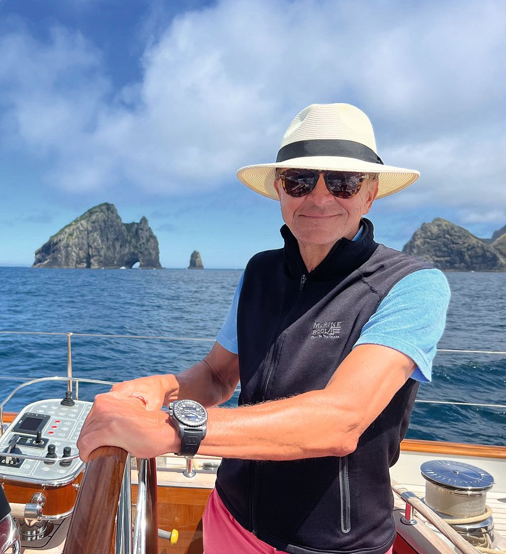 Joerg is steering the boat and smiling at the camera. He has a white panama hat on with a black band, a black gilet, blue t-shirt and pink shorts