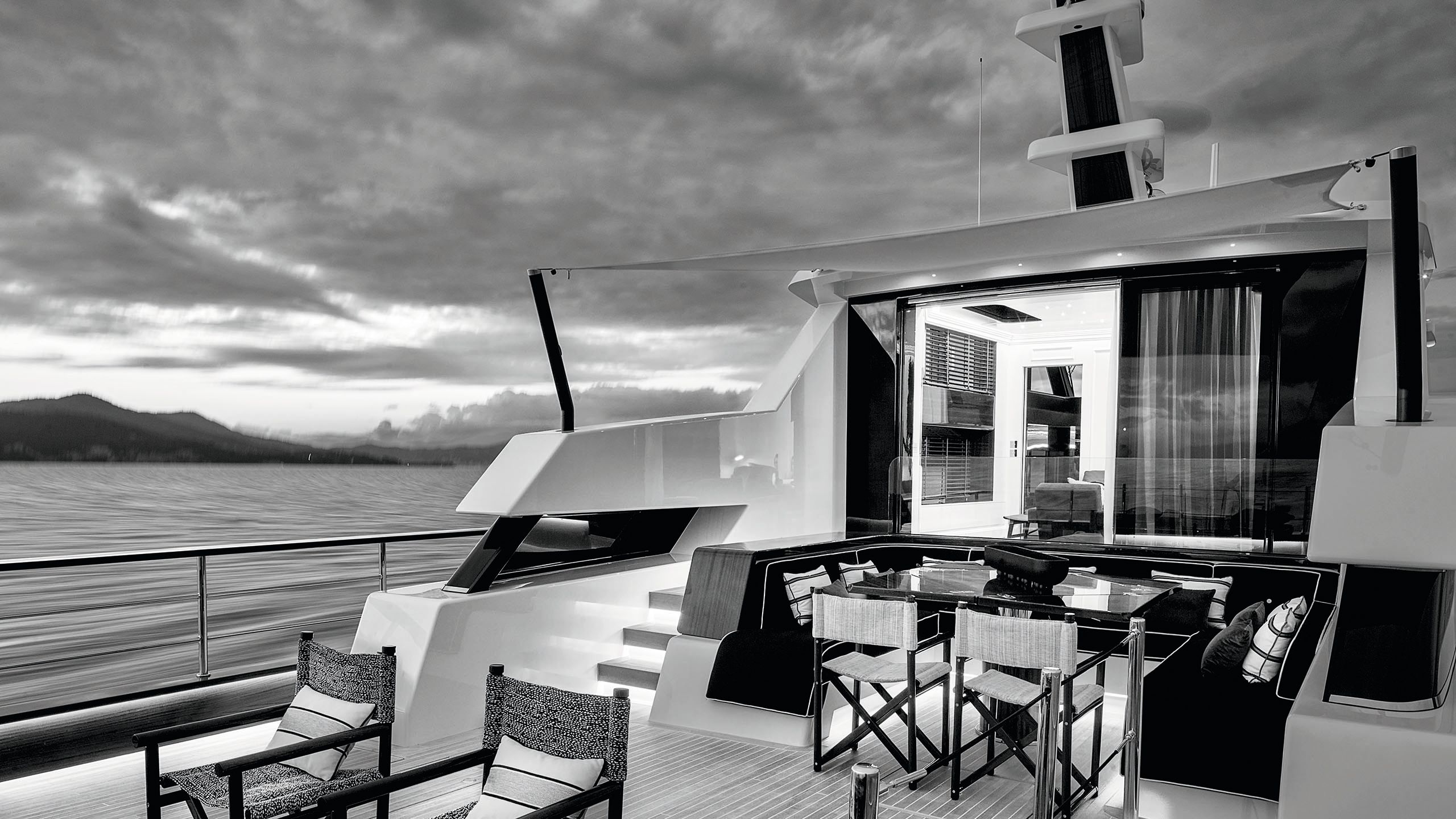 Black and white exterior shot of the deck. There are two chairs facing out on to the water and a banquette table and chairs behind for dining