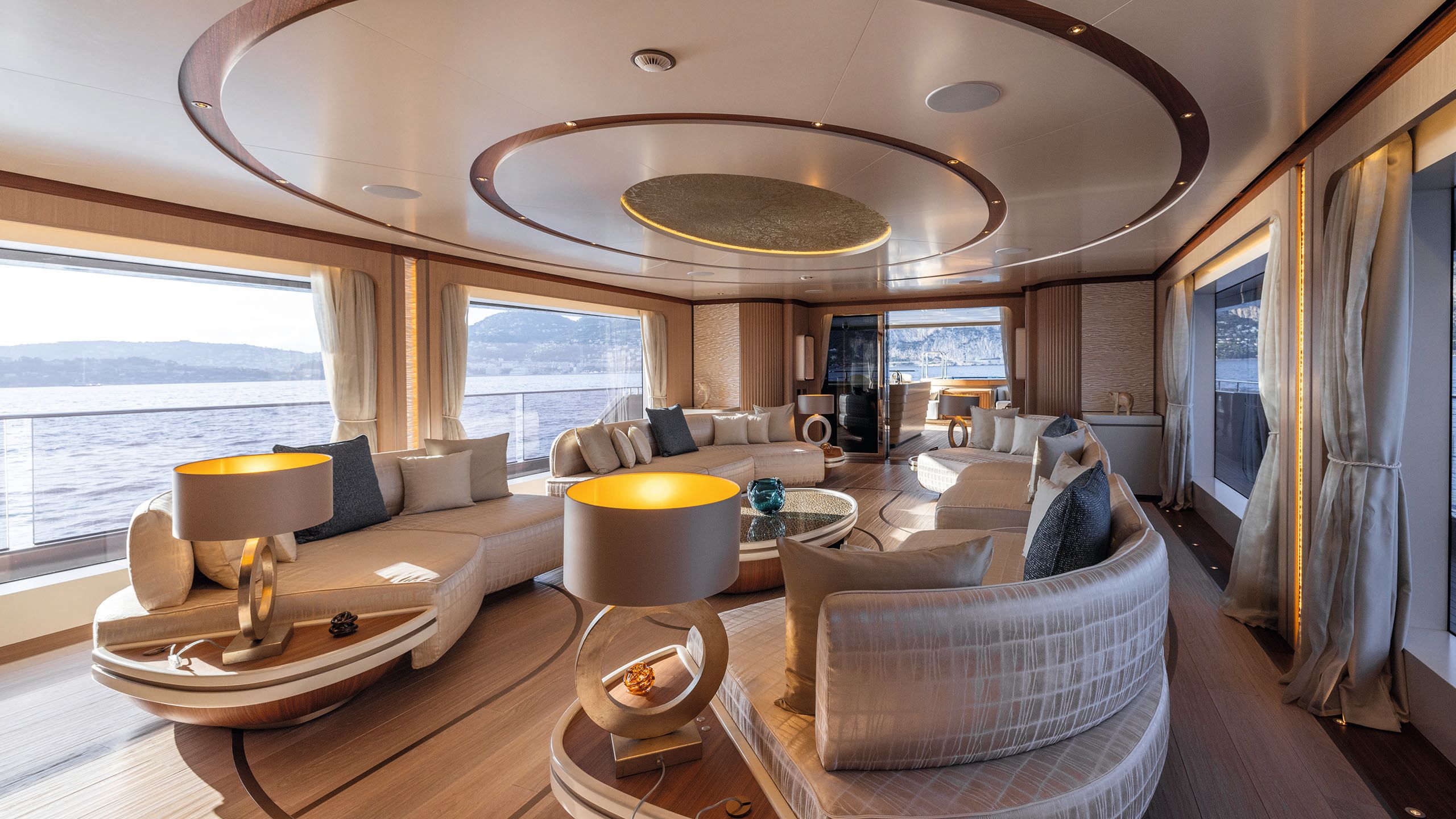 Indoor lounge with four curved sofas arranged in pairs facing each other. There is a circular motif on the ceiling and on the floor; the necks of the lamps are also circula