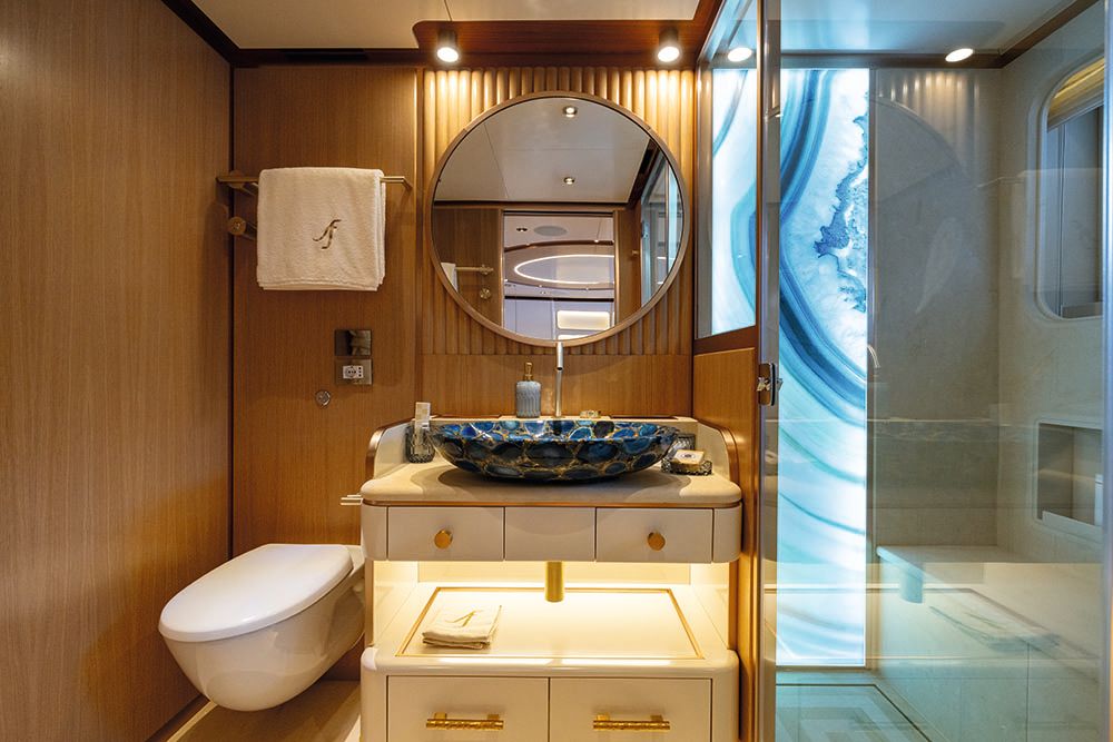 Face-on shot of a bathroom with blue sink made of gemstones. There is a circular mirror above it, a toilet to the left, and a shower to the right with a backlit blue wall