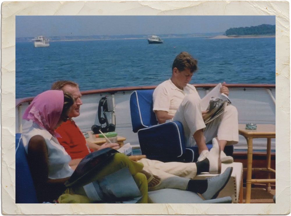 A snapshot of Kennedy with an aged cream border around it. He's sitting in the specially-designed blue chair reading a newspaper