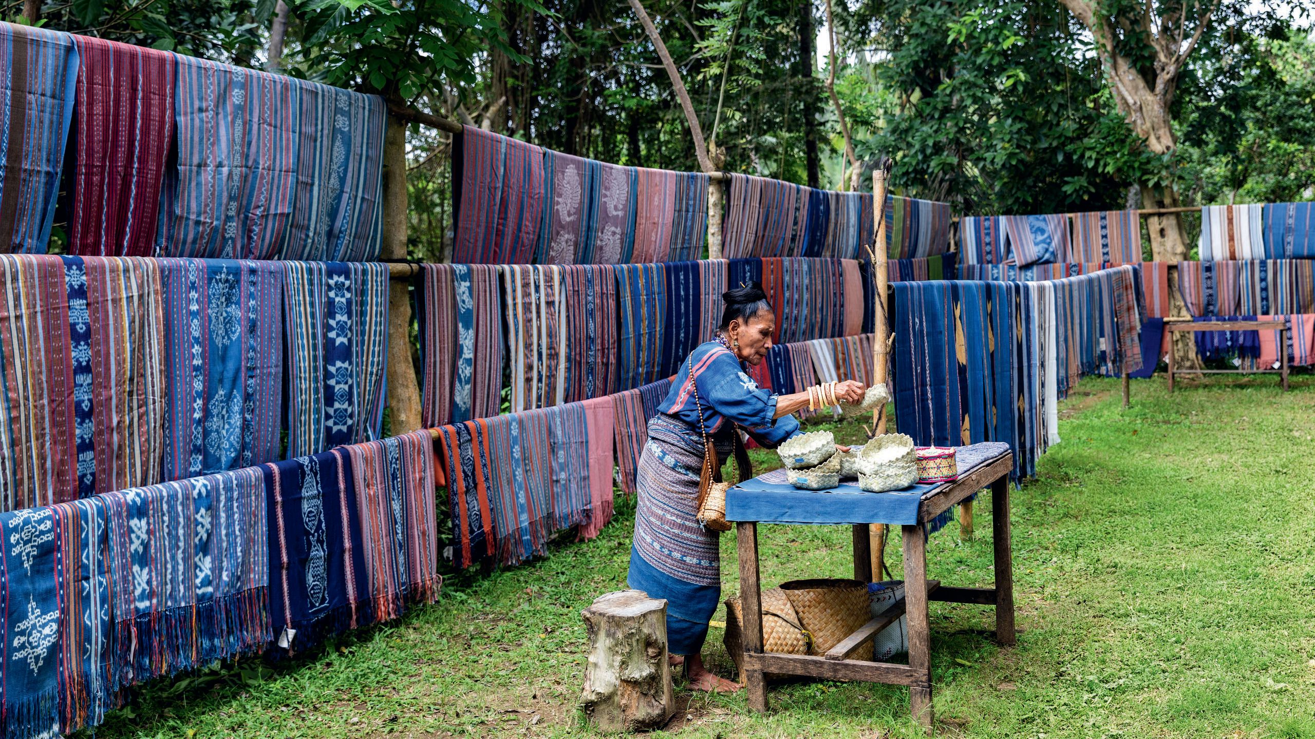 An older woman adding traditional wares to a wooden table. Behind her are rows of washing lines hung with piles of woven throws in predominantly purples, blues, and pinks