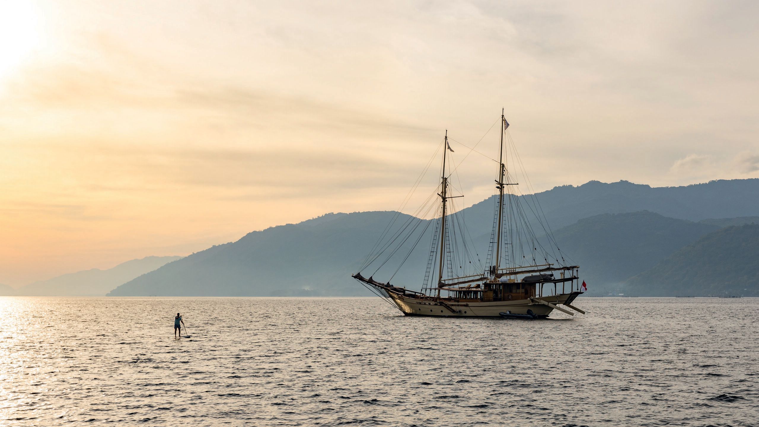 Side view of a phnisis on the water at sunrise/sunset. Its sails are down and it's almost silhouetted against background mountains. A man is on a paddleboard to the left