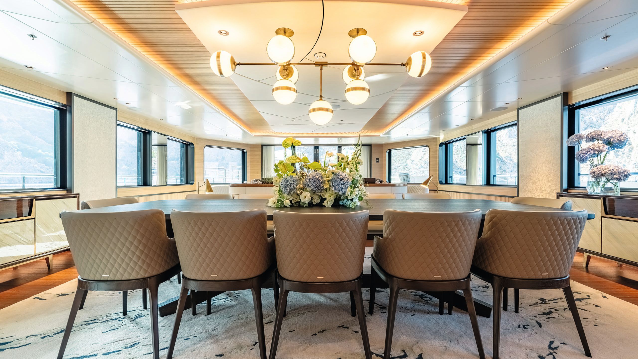 Indoor dining area with dark wood table and chairs for 12. Threre's a chandelier with globe lights and flowers in the centre of the table