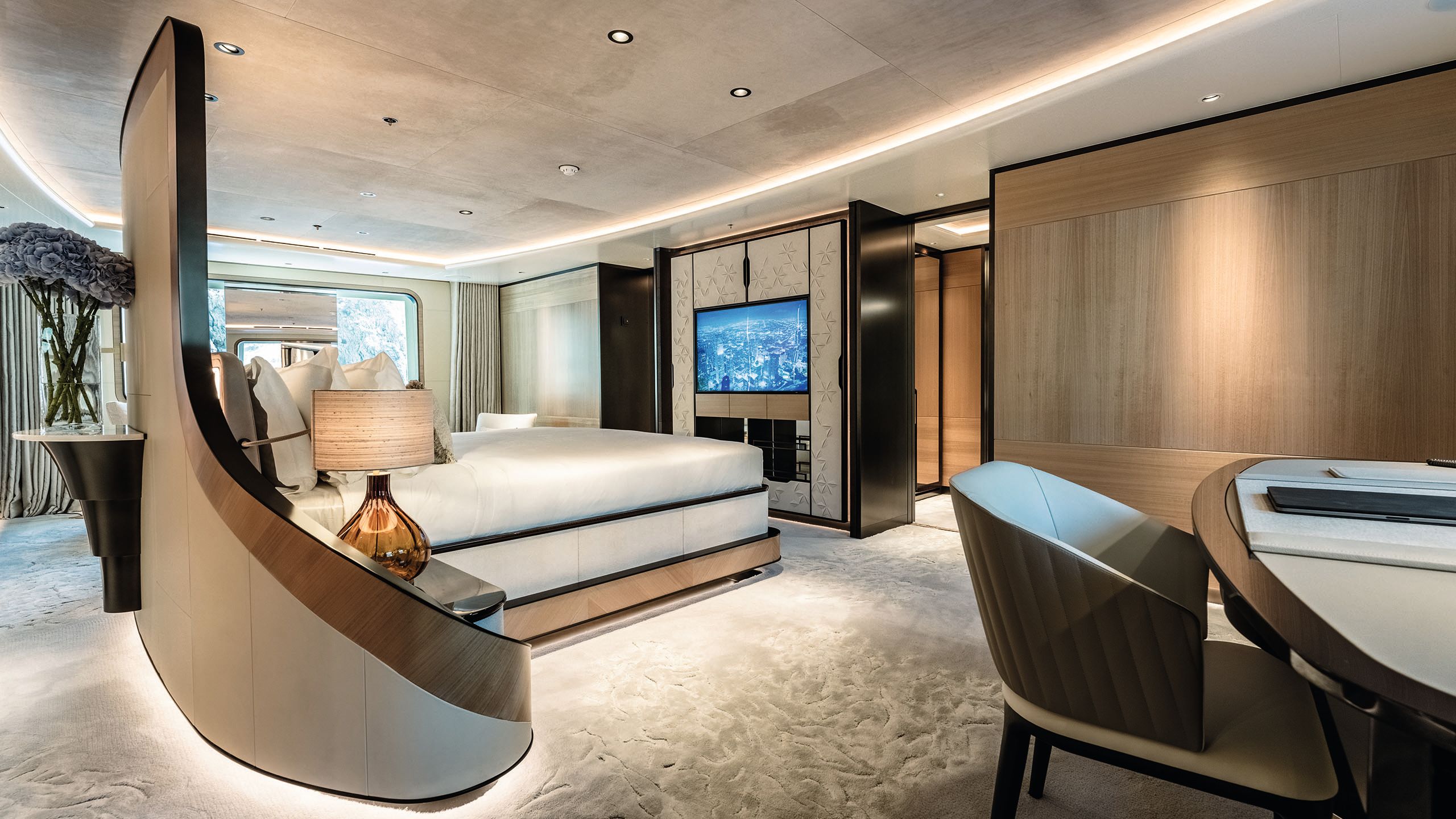Inside a large bedroom suite. There is a curved floor-to-ceiling headboard around the bed at the centre of the room, facing a TV built into cabinetry. A desk and chair are in the foreground