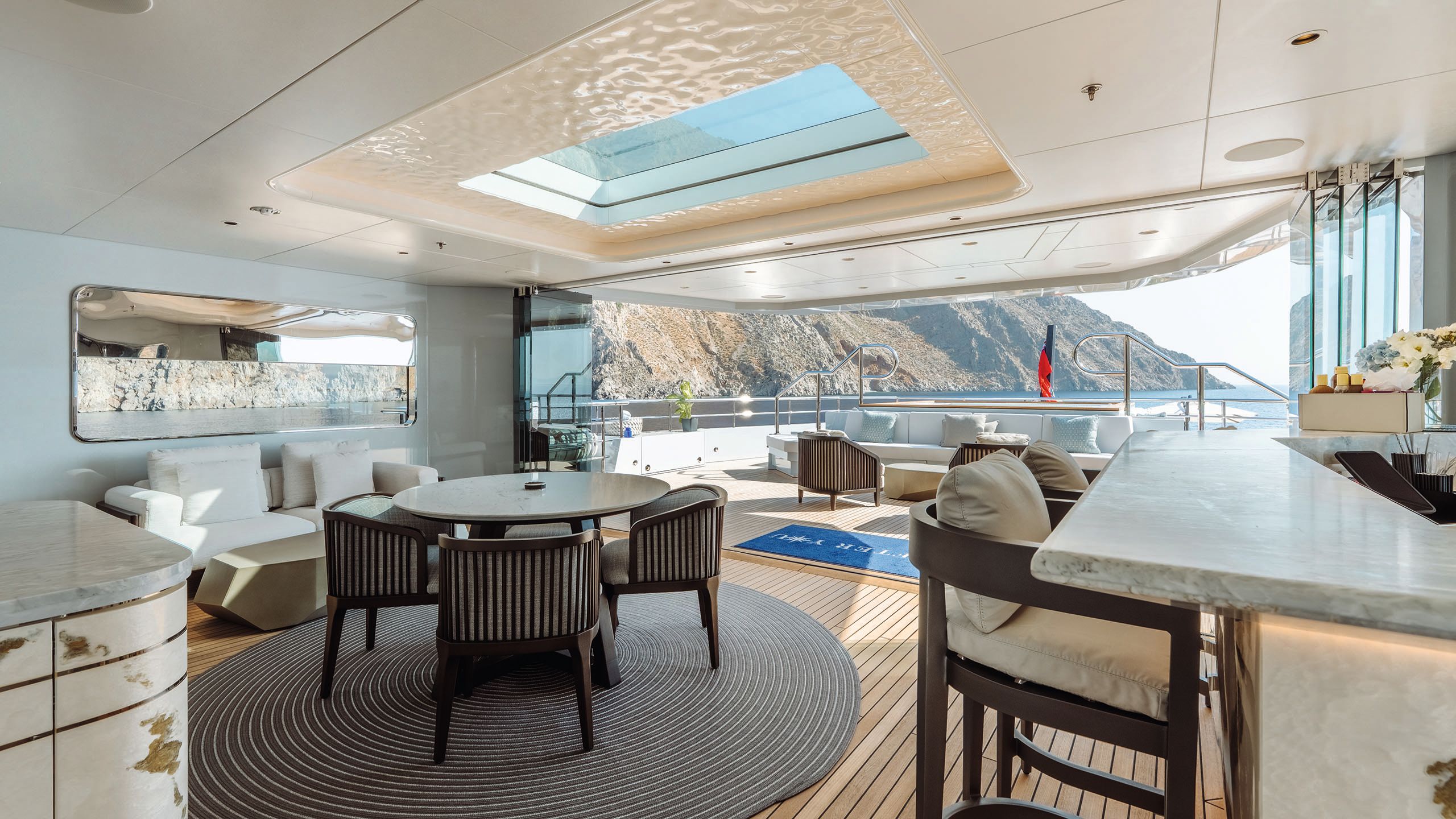 A covered dining area that opens out on to the deck. There's a small white sofa to the left and round table with seats for four in front. To the right is a counter with high chairs. In the background is soft seating on the deck