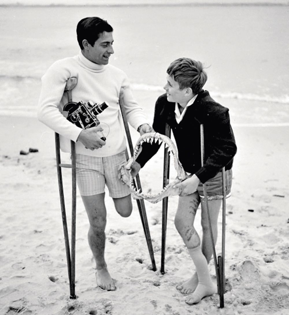 Black and white photo of Bource on crutches on the beach with a boy holding the fossilised jaw of a sea creature