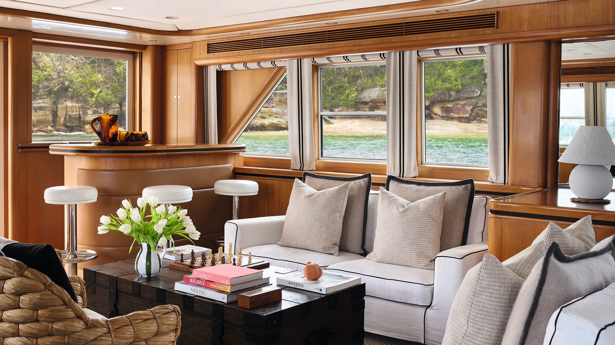 Inside Sea Raes' lounge area. There's a two seater white couch, a wicker seat opposite, a curved bar in the corner with circular white stools and a chess set, books and flowers on the chest-style coffee table