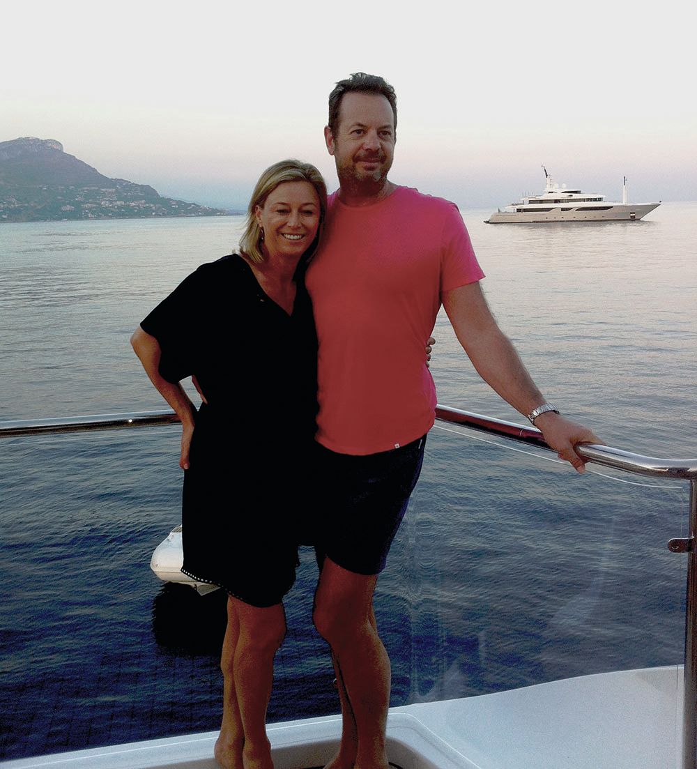 Rockman has his arm around his wife. They're pictured on a boat and he's wearing a pink t-shirt and shorts and she's wearing a black dress