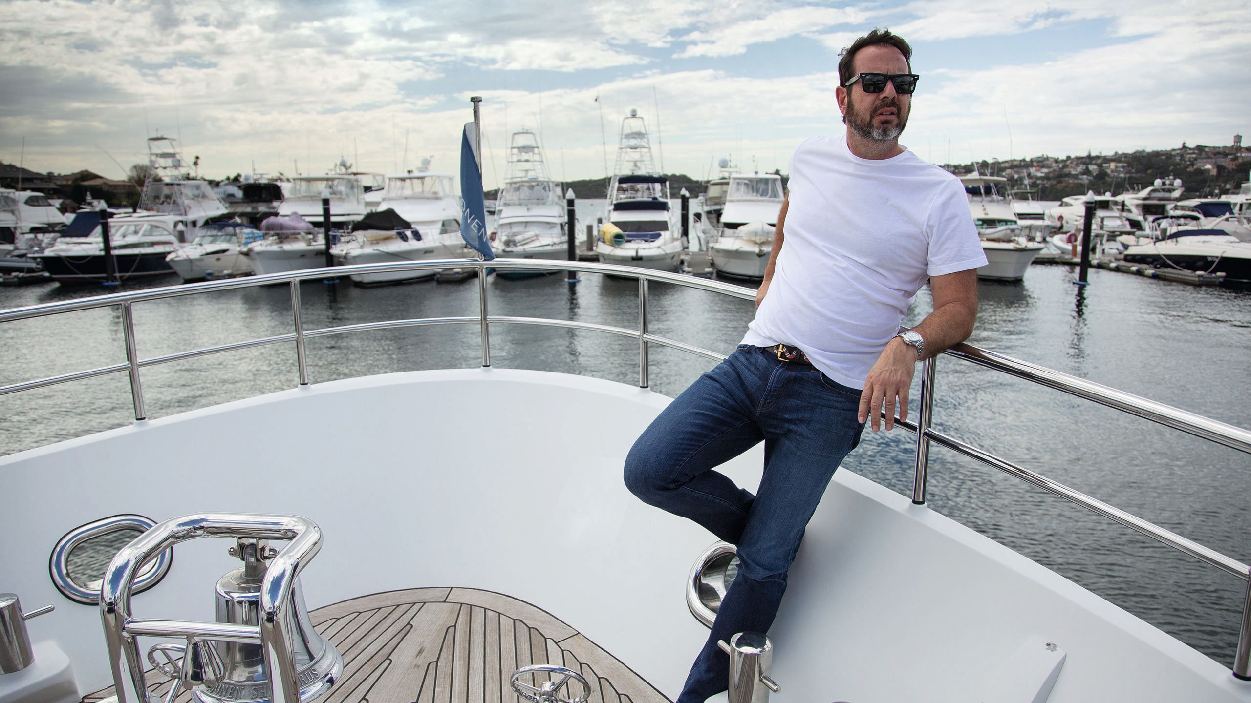Rockman leaning on the railings at the nose of the boat. He's wearing a white t-shirt, dark jeans and sunglasses. A harbour filled with boats is behind him