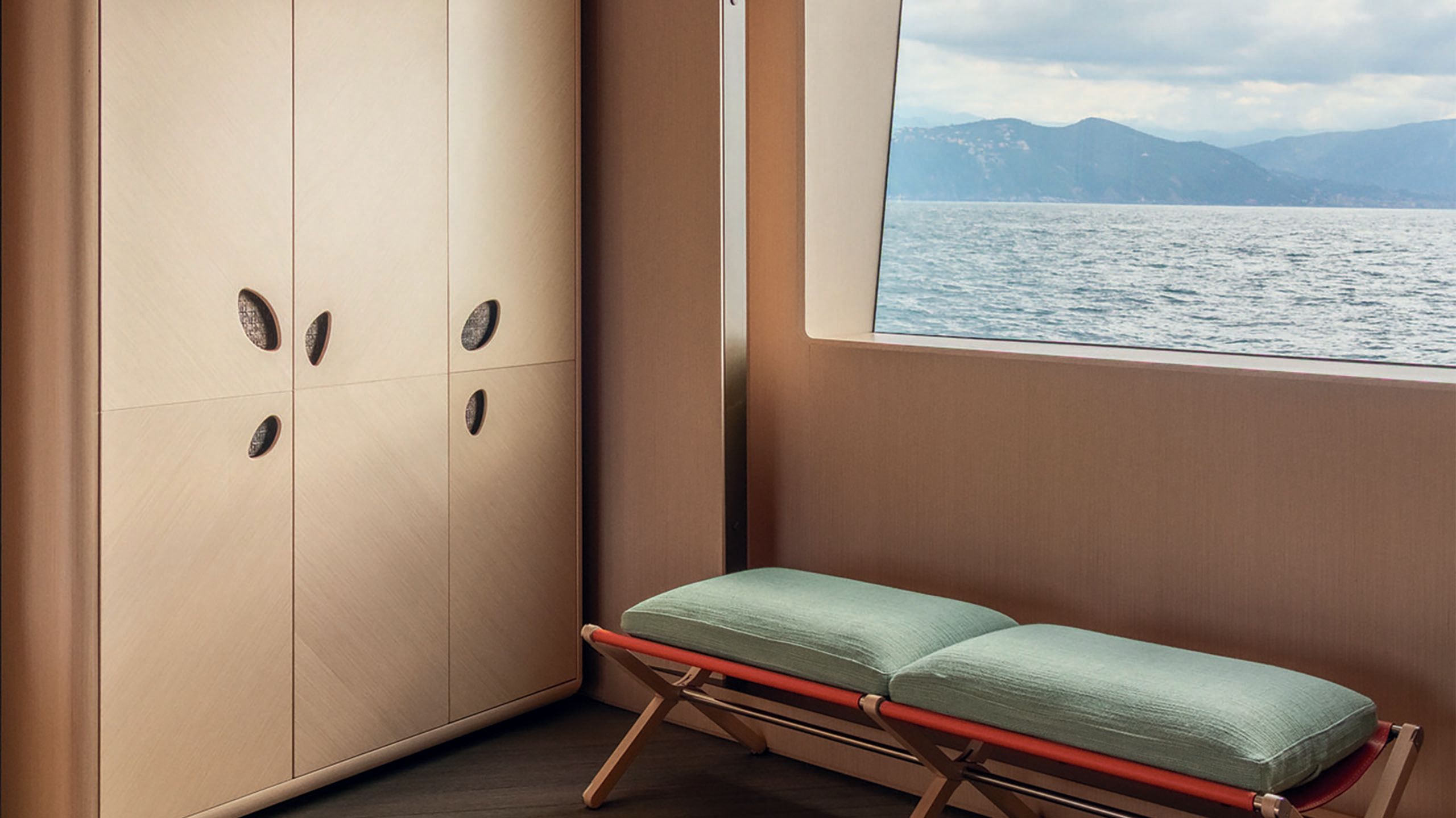 A padded bench with green cushions next to a rectangular window looking out on to the ocean