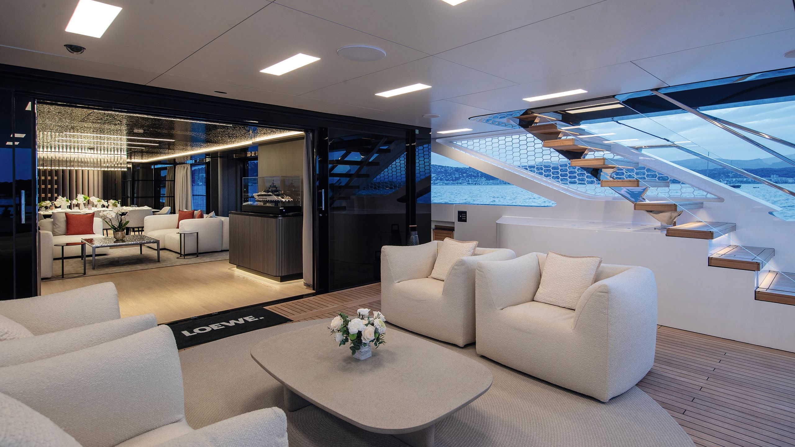 A covered outdoor deck area that leads into the main saloon. There is loose off-white armchairs and a neutral coffee table between them