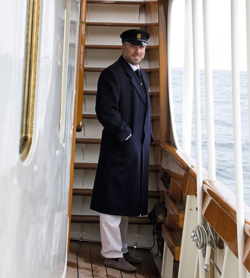 Colour photo of a man standing on a deck in front of a staircase. He's wearing a long navy coat, navy hat and white shirts