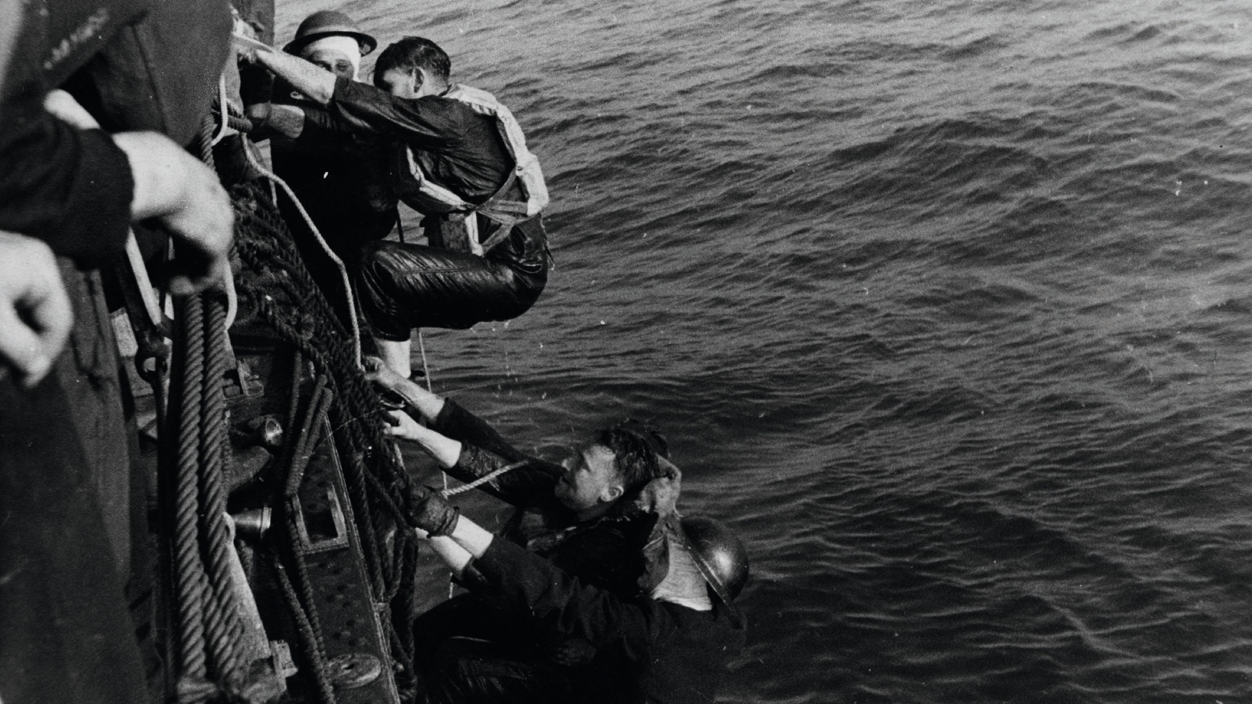 Black and white photo of troops climbing up ropes on the side of a vessel