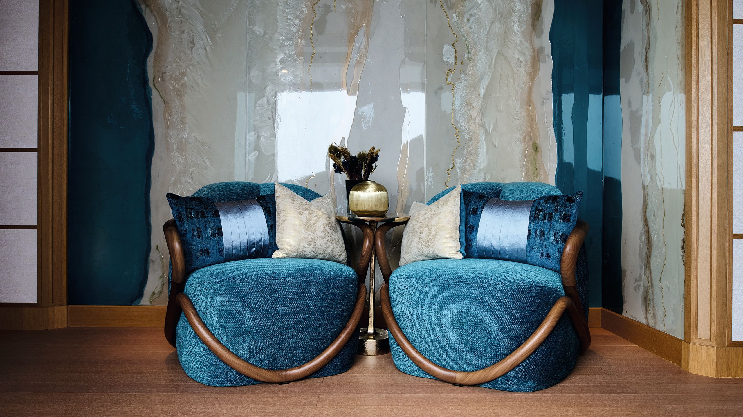 Two jewel-toned blue chairs with walnut wood finish on the arms. They have blue and gold cushions and a gold table between the arms which has a gold vase with dried flowers in. The wall behind has an inlay effect