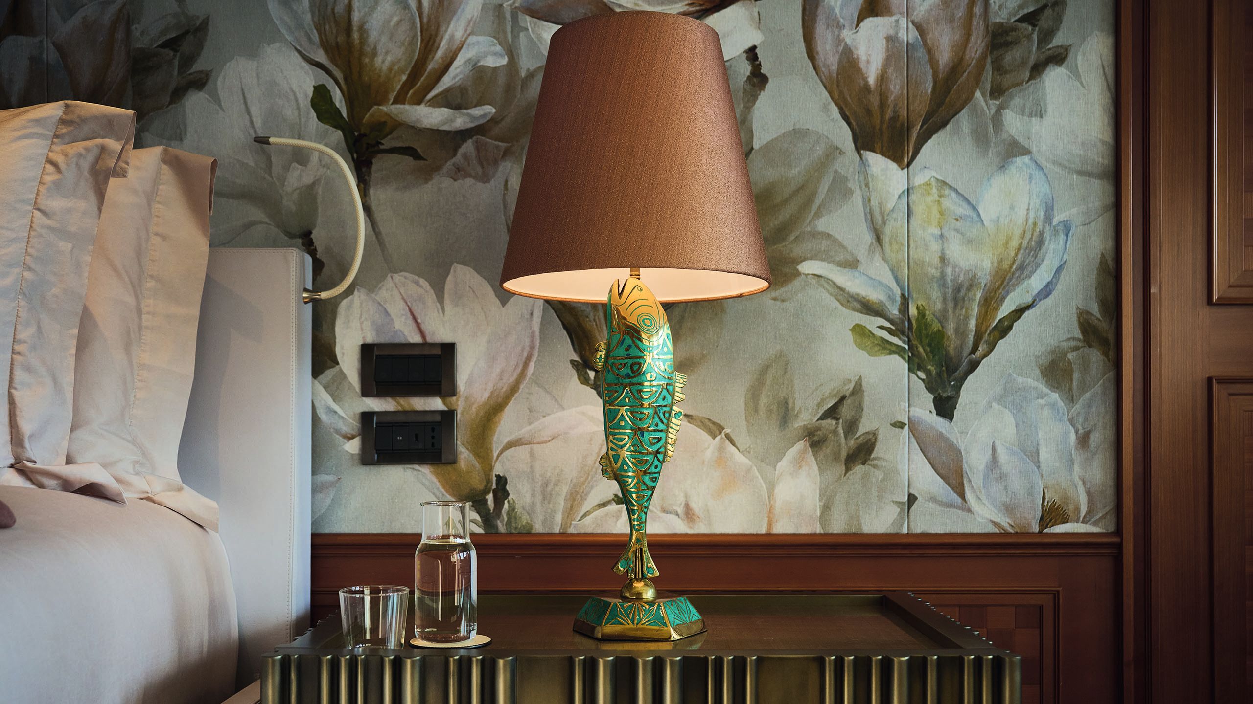 A close-up of a beside table lamp. The stand is a gold and turquoise fish and the shade is a dusky pink