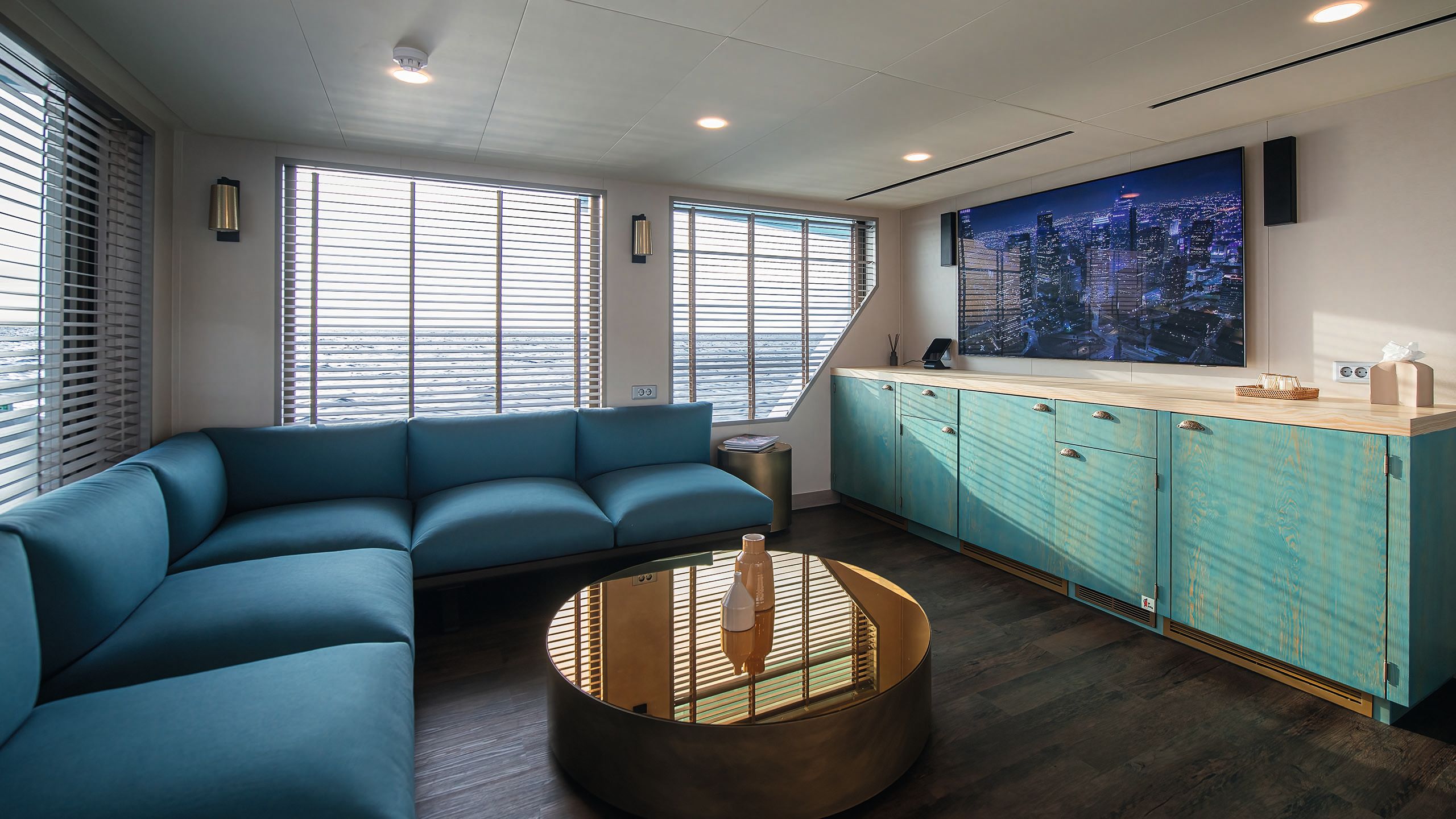 A small turquoise L-shaped sofa with round table in front with metallic top. They face a wall-mounted TV above an aquamarine cabinet