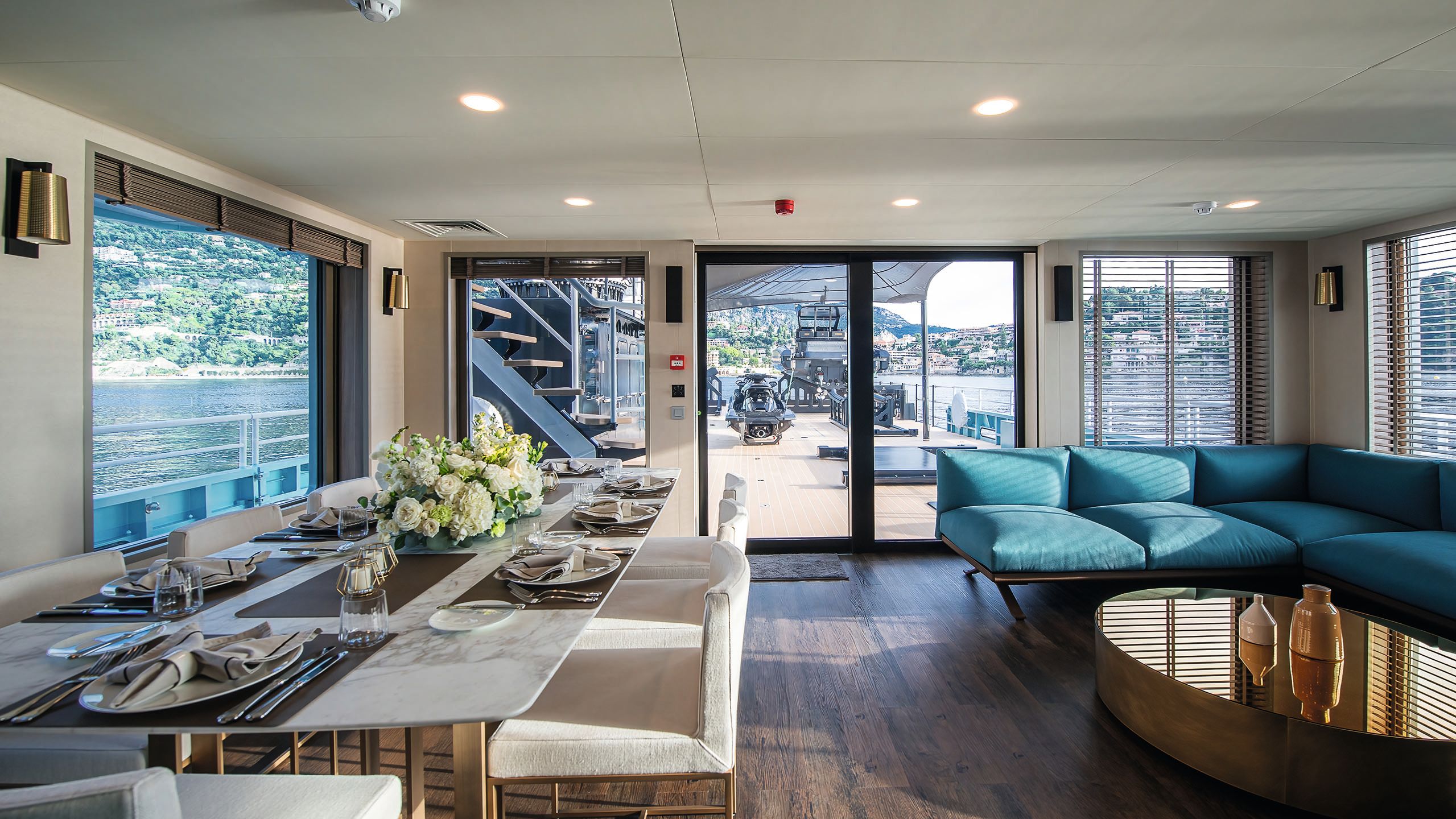 A marble-effect dining table on the left which place settings. A turquoise L-shaped sofa is on the right with a large oval table with metallic top in front