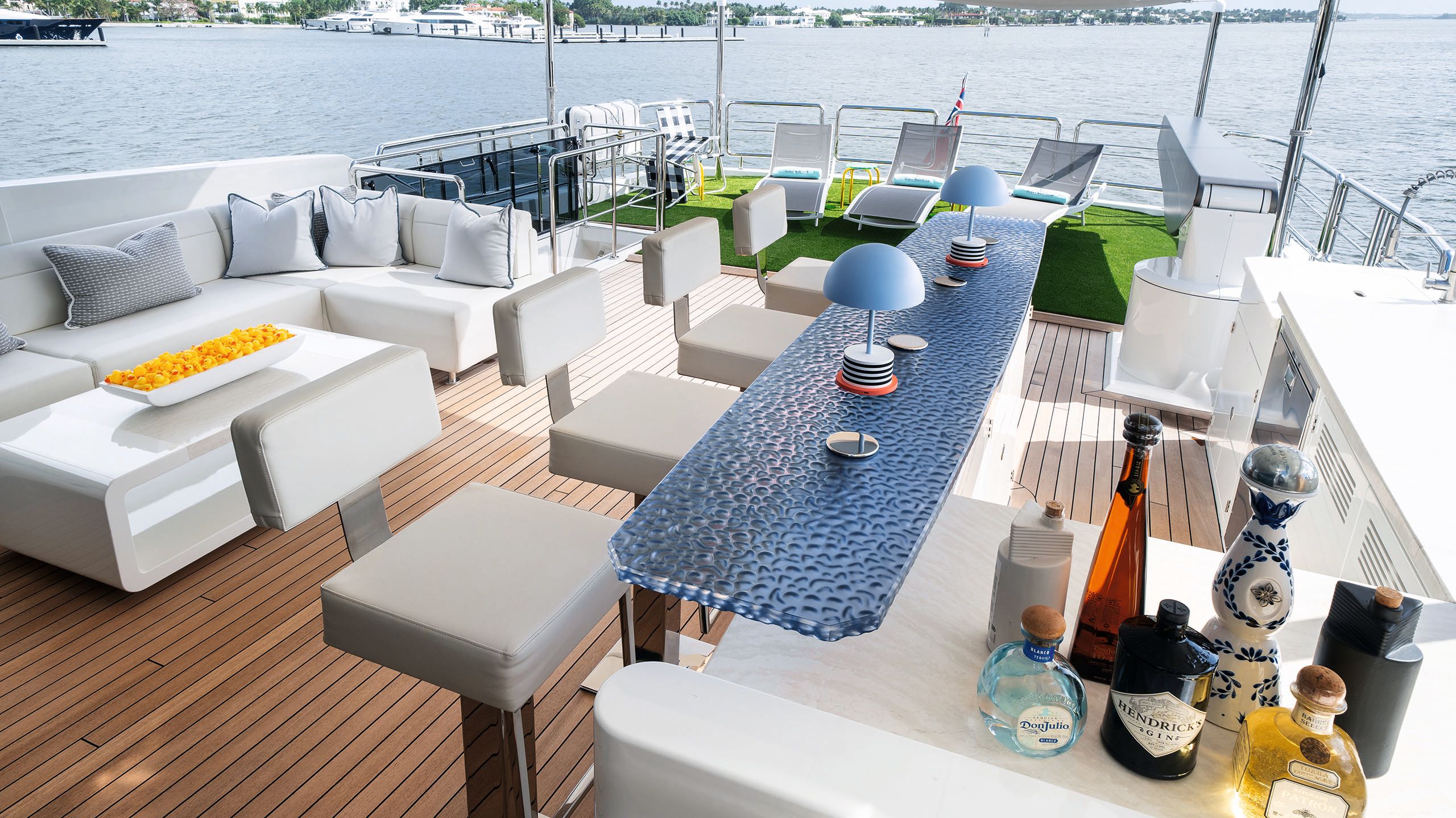 Deck area with bar stocked with various spirits and a blue glass countertop. There are high stools in front of it and behind an L-shaped sofa with a white table holding a dish full of plastic yellow ducks. At the back of the deck is a section of AstroTurf and three deckchairs
