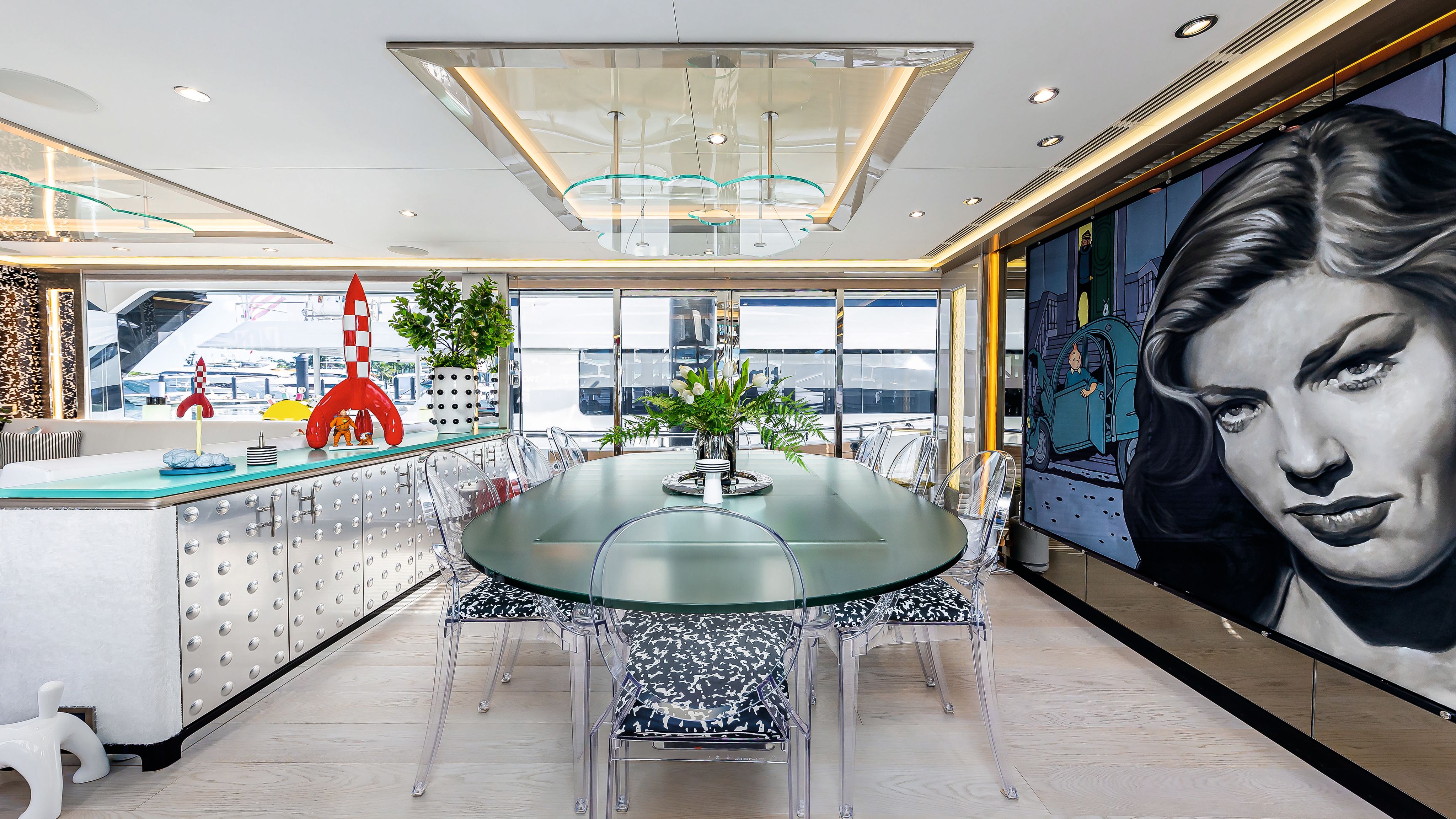 A dining room with clear perspex frosted table and matching chairs with black and white cushions that have an abstract print. To the left is a long sideboard with silver doors and a red and white checked rocketship on the top. To the right is a huge mural of TinTin with a black and white image of Lauren Bacall