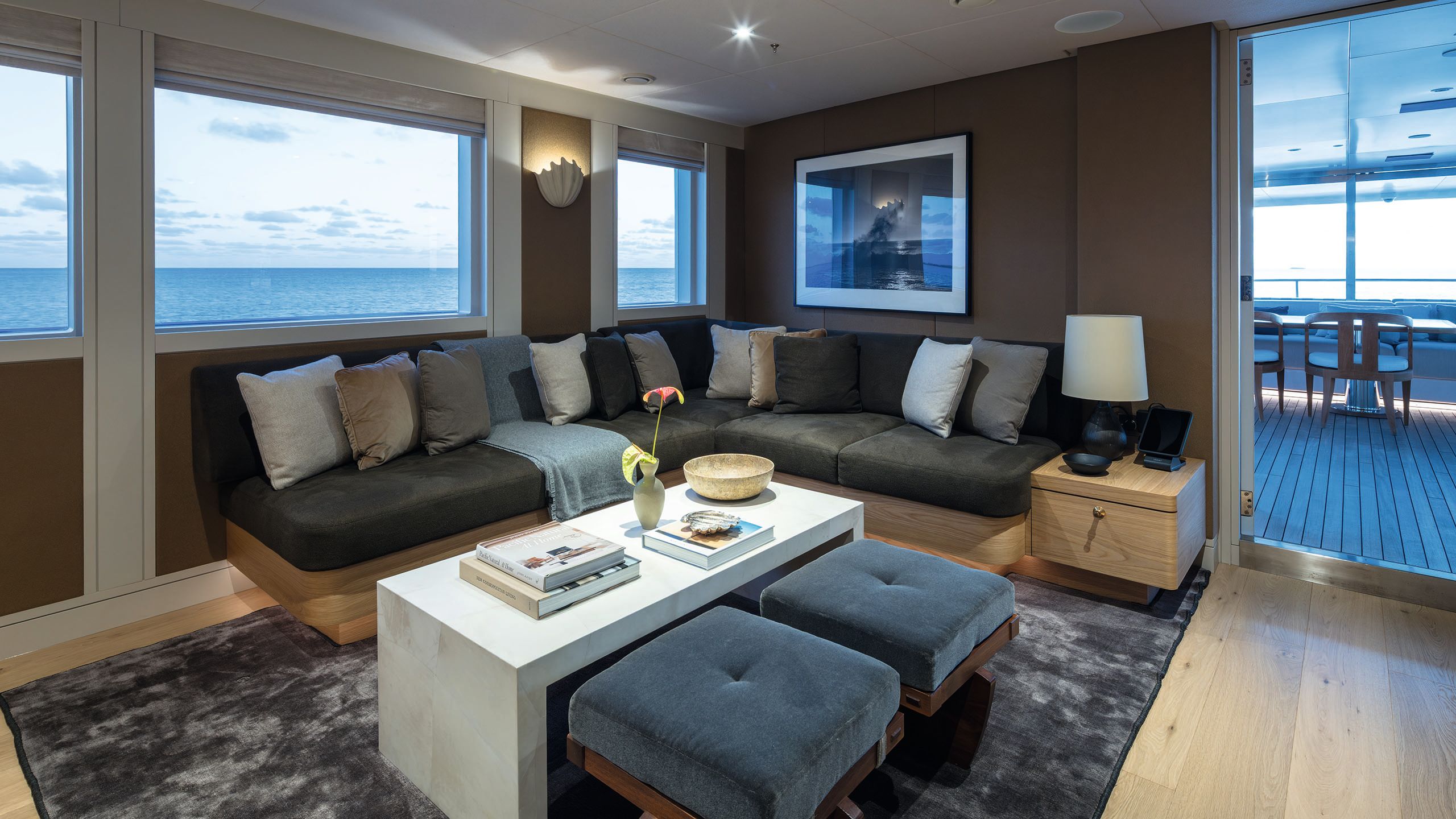 Upper deck saloon with L-shaped sofa in dark grey with throw cushions in blue, grey and white. There is a white rectangular table in front with two blue footstools