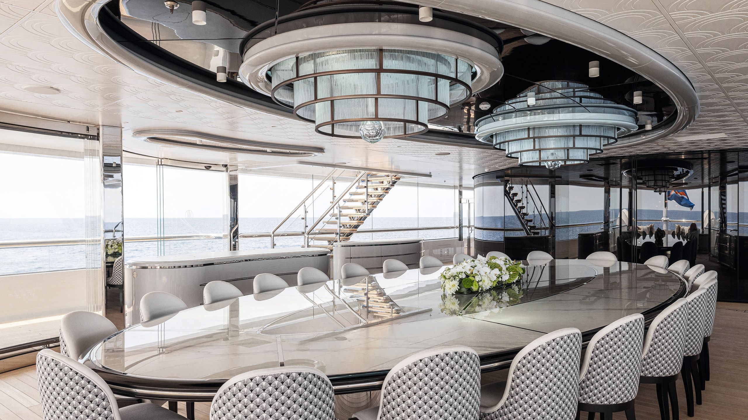 Curved dining tabled in neutral colour with glossy top. There are seats for 25 which have a black and white repeating fan pattern on the back. Above are two tiered chandeliers