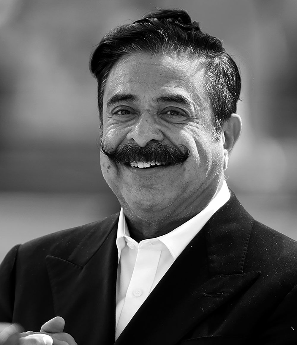 Black and white image of Khan's father. He is smiling at the camera and clasping his hands. He's wearing a white shirt and black suit jacket